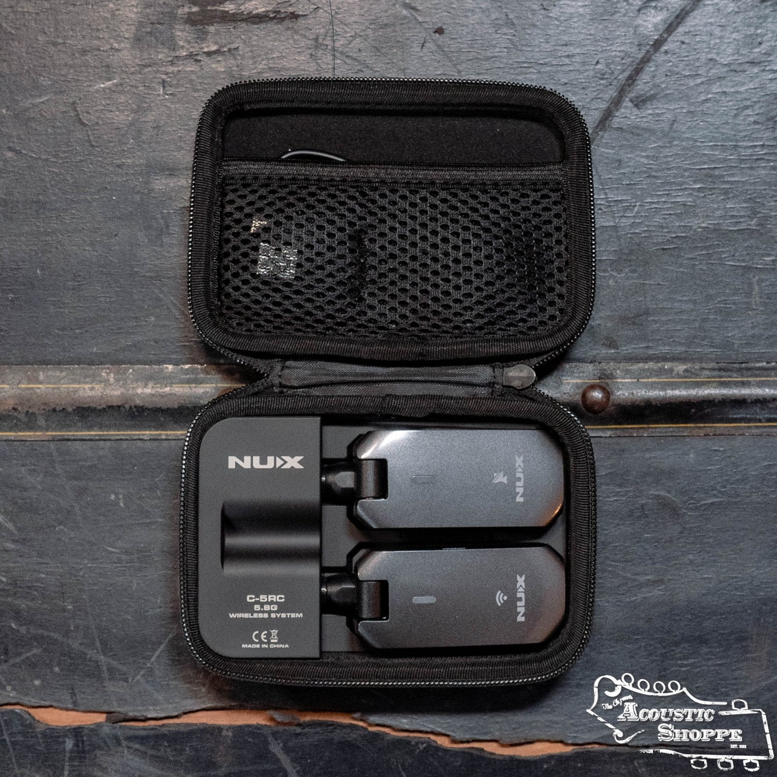 A small black zippered case rests open on wood, showing the NUX C-5RC 5.8GHz Wireless Guitar System for Acoustic or Electric Guitar (Black) by NUX with two black units and a mesh pocket inside. The Acoustic Shoppe logo appears in the lower right corner.