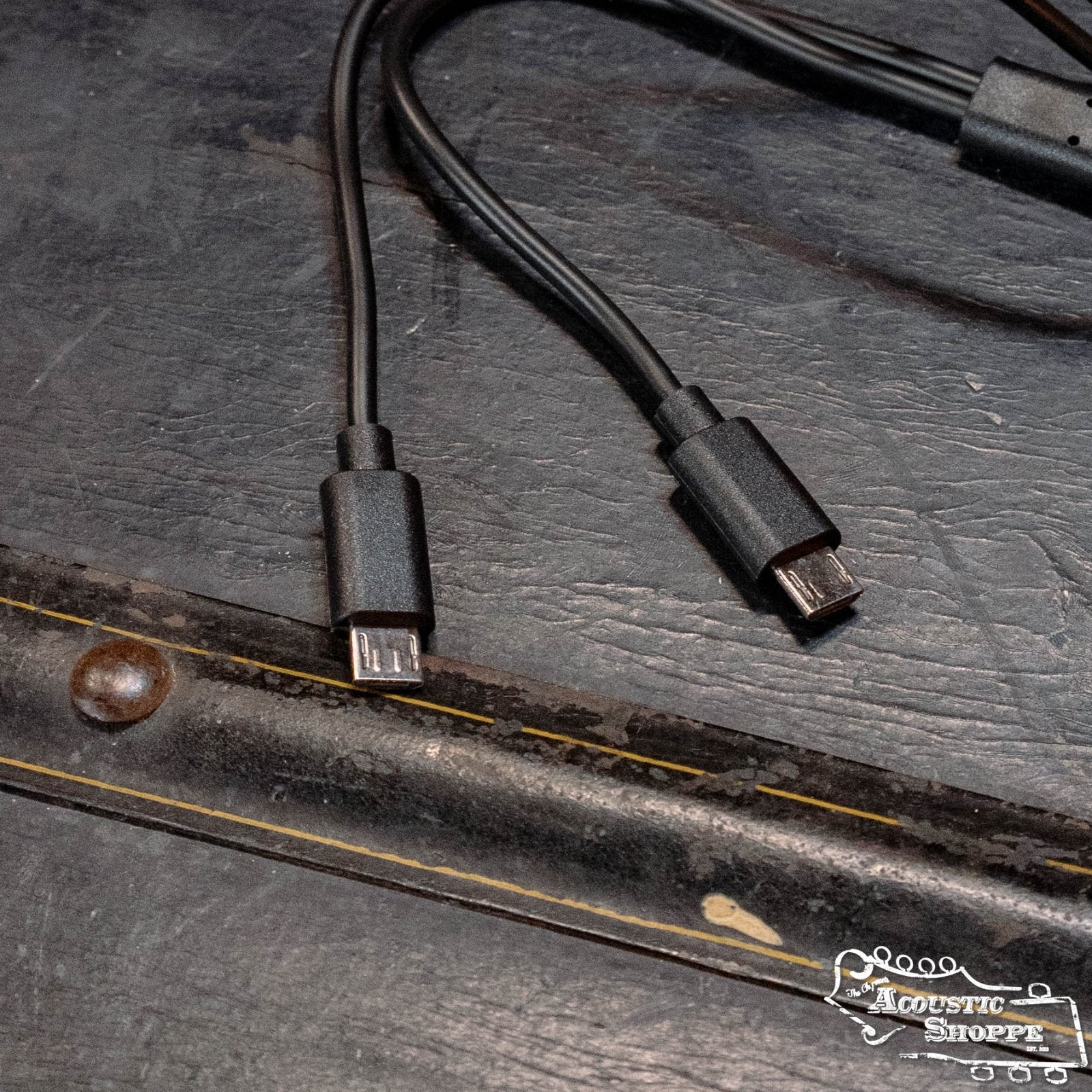 Two black USB cables with micro USB and USB-C connectors lie on a dark surface near a metal edge—ideal for your NUX C-5RC 5.8GHz Wireless Guitar System for Acoustic or Electric Guitar (Black). The Acoustic Shoppe logo is in the bottom right.