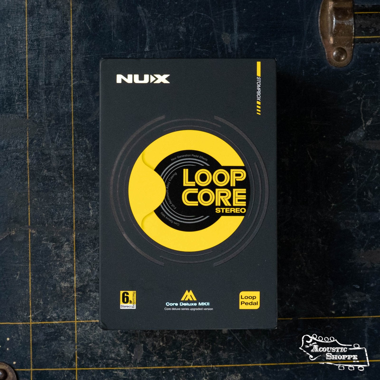 Top-down view of the NUX Loop Core Stereo guitar pedal from NUX, shown in its box with bold graphics. This stereo looper features drum patterns and MIDI compatibility, and rests on a dark grid-patterned surface.