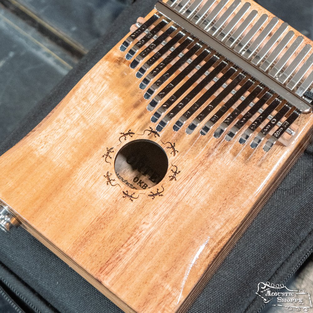 A close-up of the Ortega OKB1E Lizard Series Solid Acacia Wood Kalimba with 17 keys, built-in pickup, and gecko designs around the sound hole, resting on a dark fabric surface.