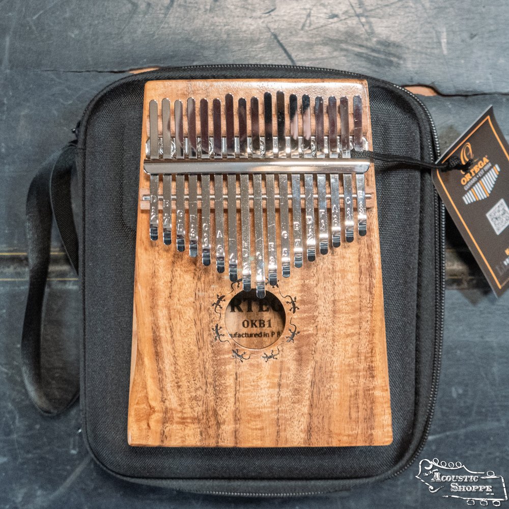 The Ortega OKB1 Lizard Series Kalimba features 17 metal tines, a solid acacia wood body with a natural finish, decorative carvings around the sound hole, and comes with a black carrying case tagged with the Ortega brand.
