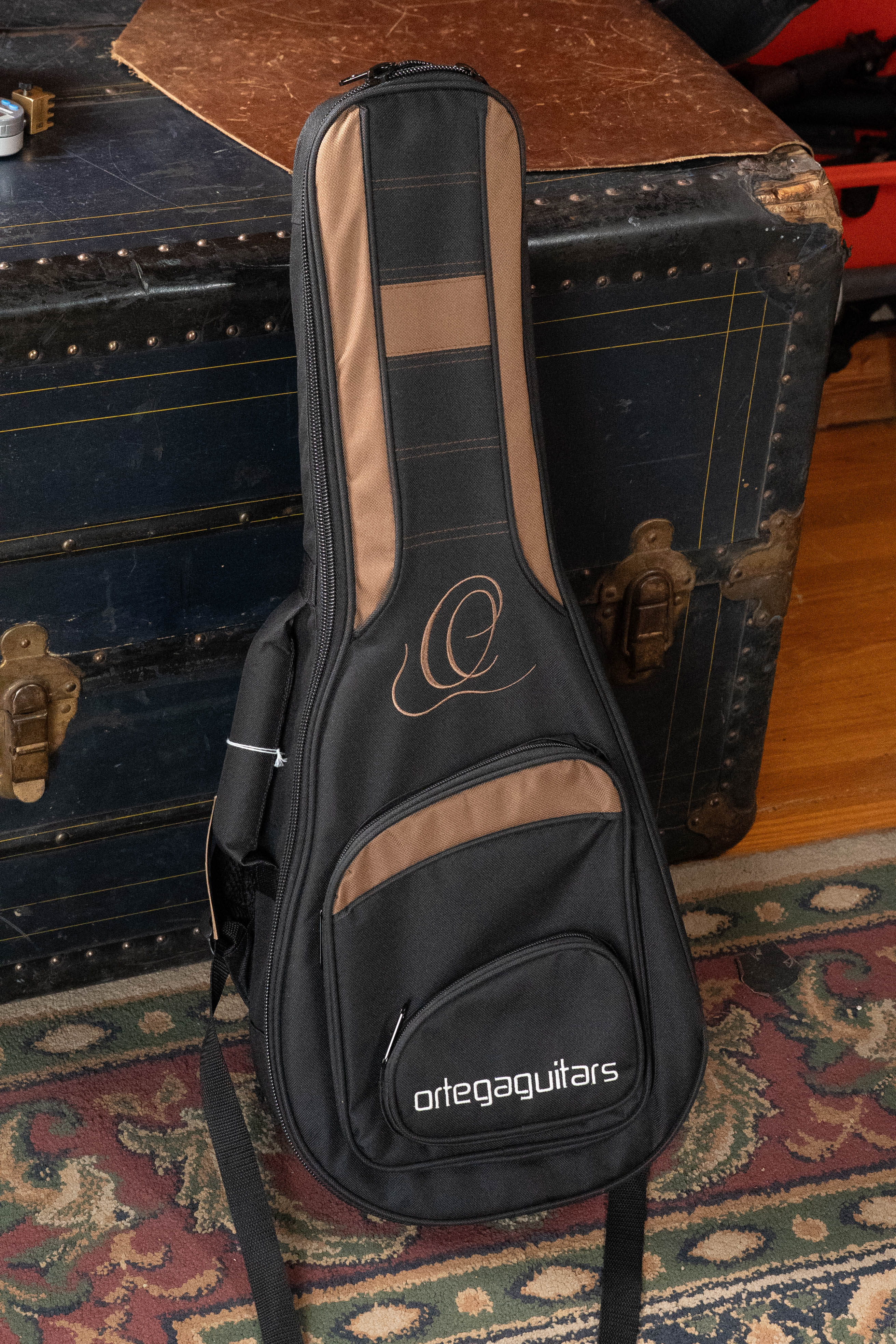 An Ortega ONB-MA Standard Size Mandolin Bag in brown with black stands upright on a patterned rug in front of a vintage trunk with brass hardware.