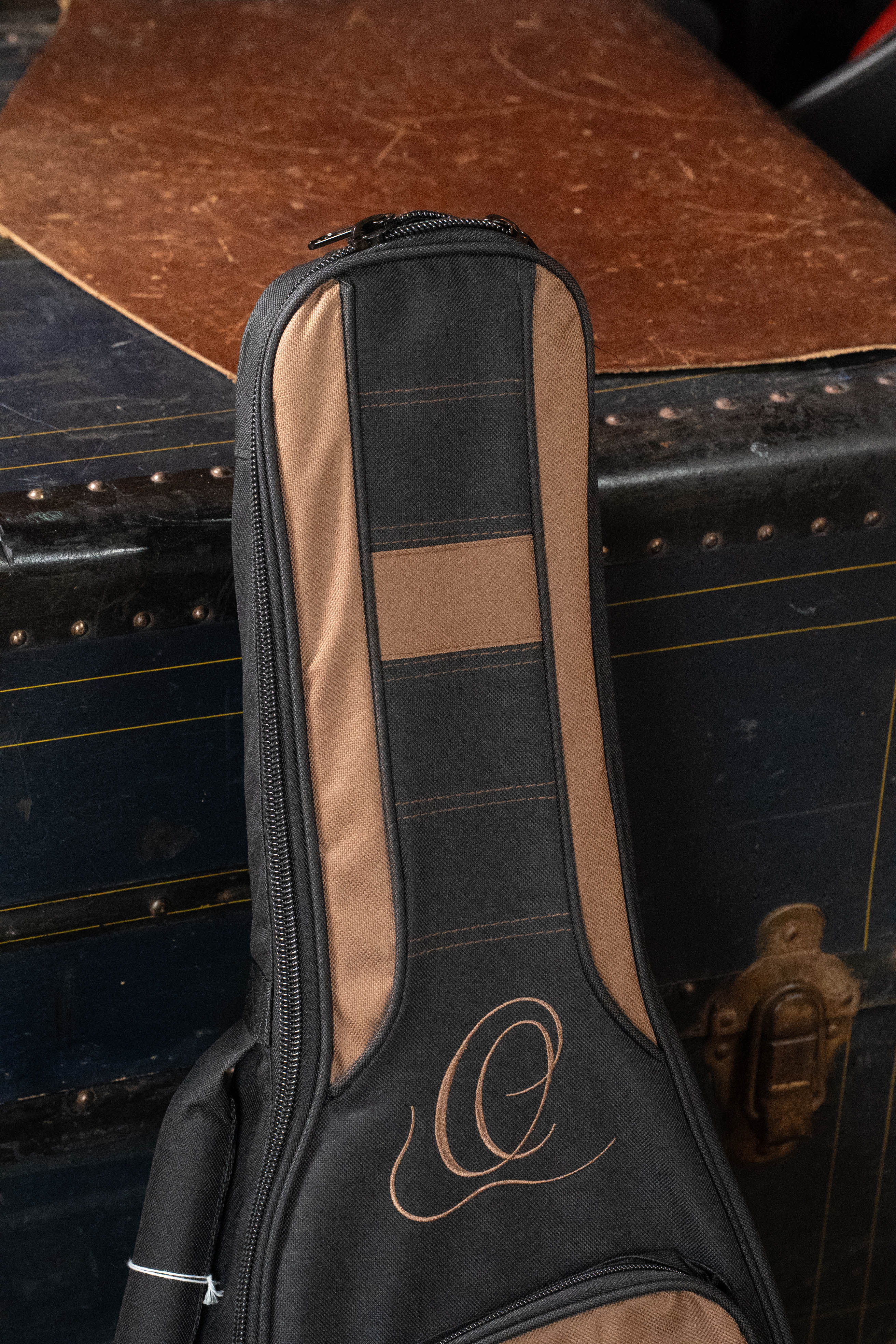 The Ortega ONB-MA Standard Size Mandolin Bag in brown with black stands upright before a vintage, dark blue trunk featuring brass hardware and a worn brown top.