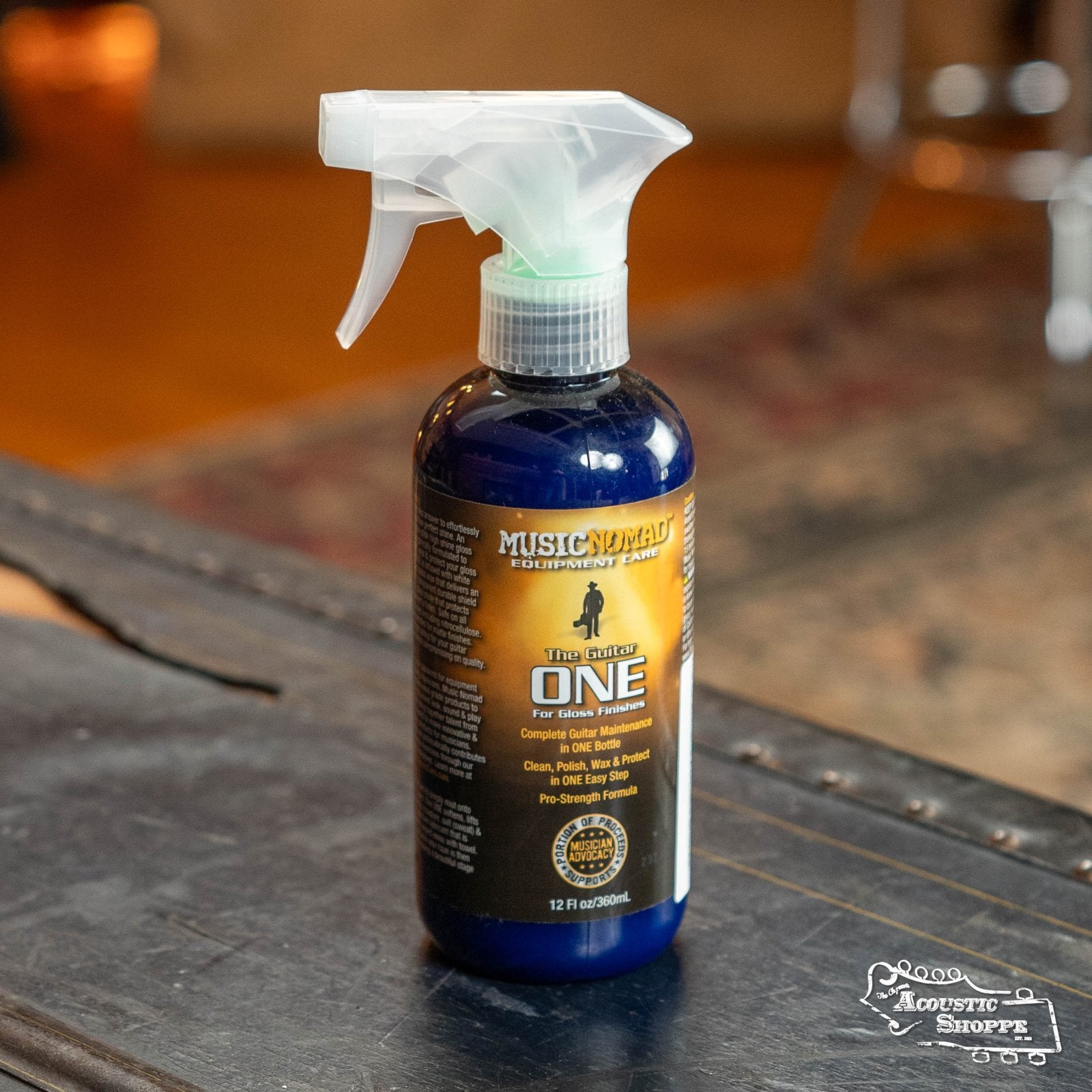 A bottle of Music Nomad The Guitar One 12 oz. Tech Size (MN150) All in 1 Cleaner, Polish & Wax sits on a wooden table, featuring its carnauba wax formula. The Acoustic Shoppe logo and a blurred indoor room are visible in the background.