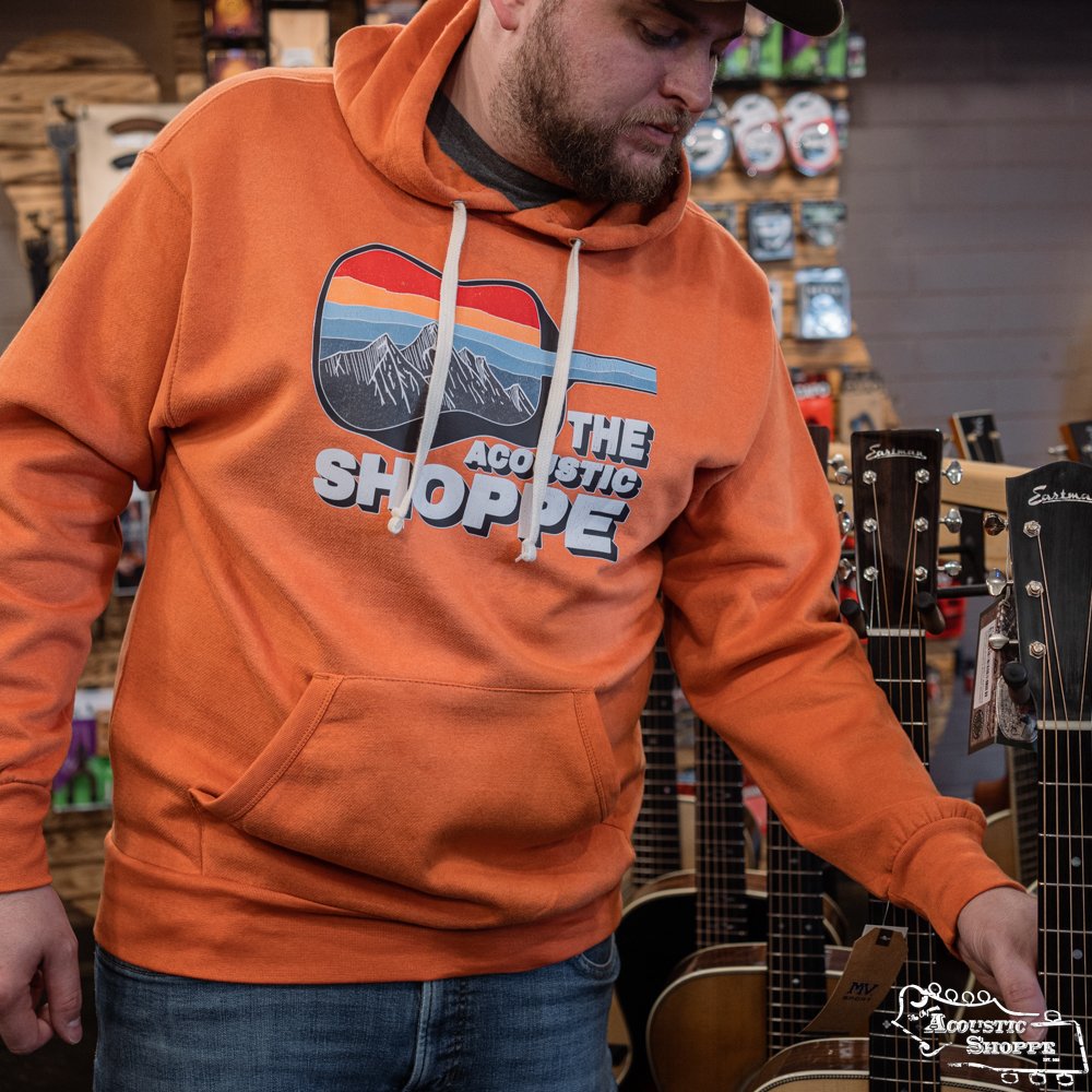 A man wearing an Amplify Screen Printing & Promotions TAS Scenic Guitar MV Sport Vintage Fleece Hoodie in orange stands among acoustic guitars in a music store, reaching for one of the instruments.