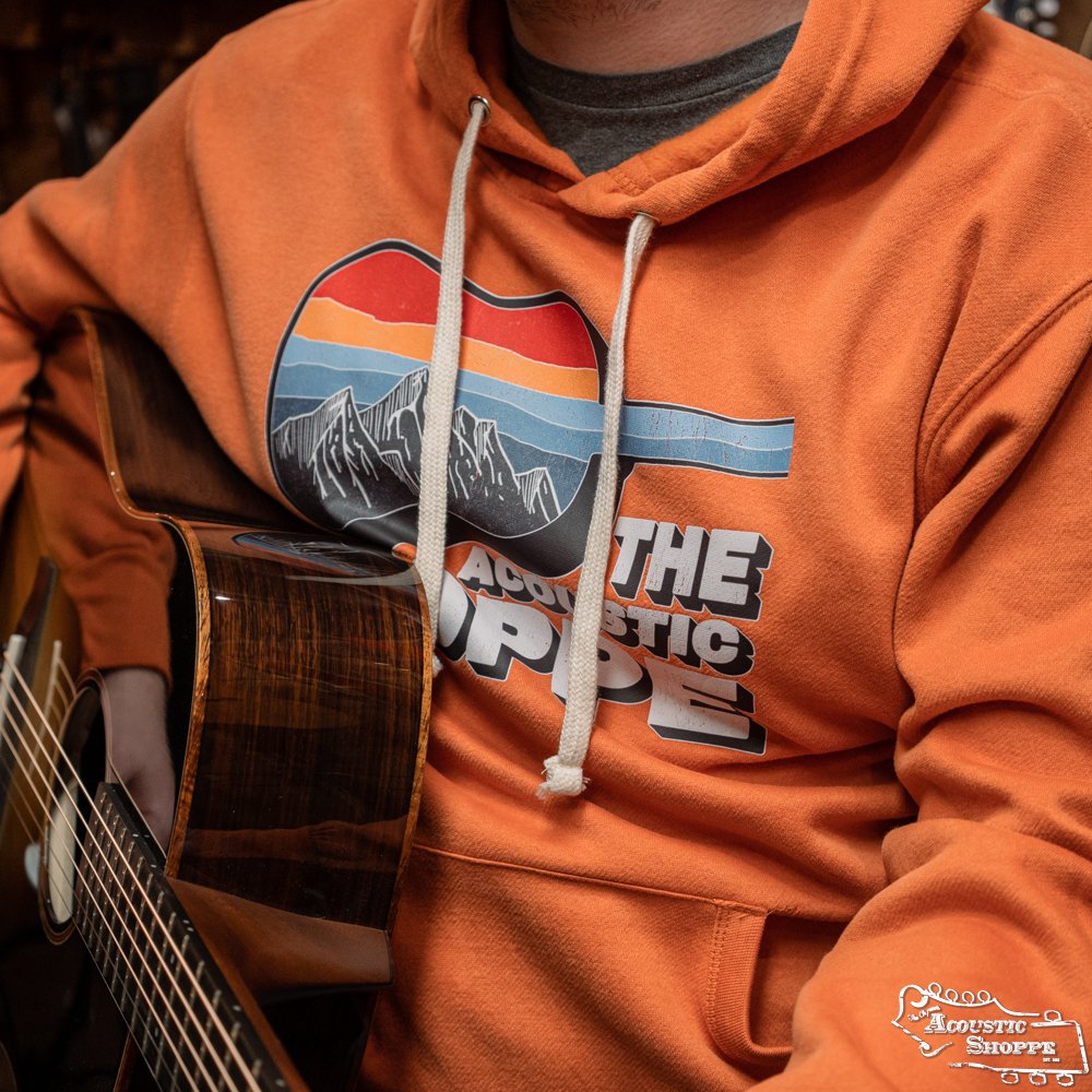 A person in the TAS Scenic Guitar MV Sport Vintage Fleece Hoodie - Orange by Amplify Screen Printing & Promotions, featuring a mountain and sunset graphic and "The Acoustic Shoppe" text, plays an acoustic guitar, showing only their upper body and guitar.
