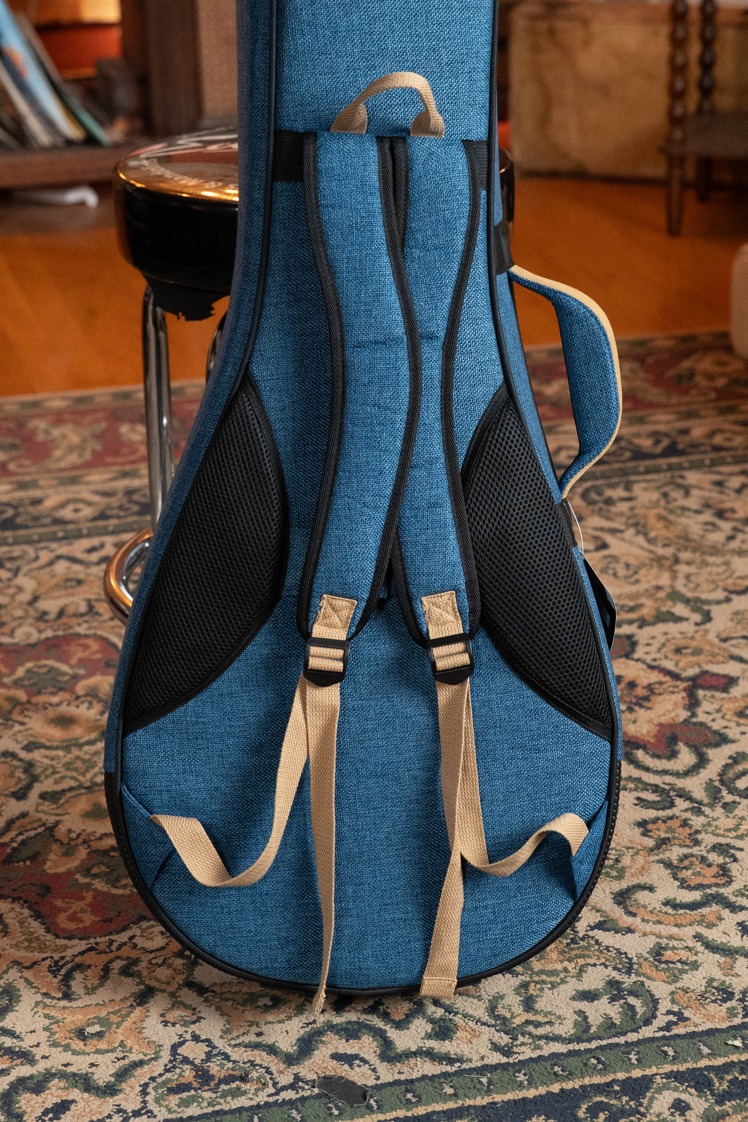 The Ortega OSOCABJ-OC Banjo Soft Case in ocean blue features beige backpack straps and black mesh pockets. It stands upright on a patterned rug, with a metal stool and other furniture seen in the background.