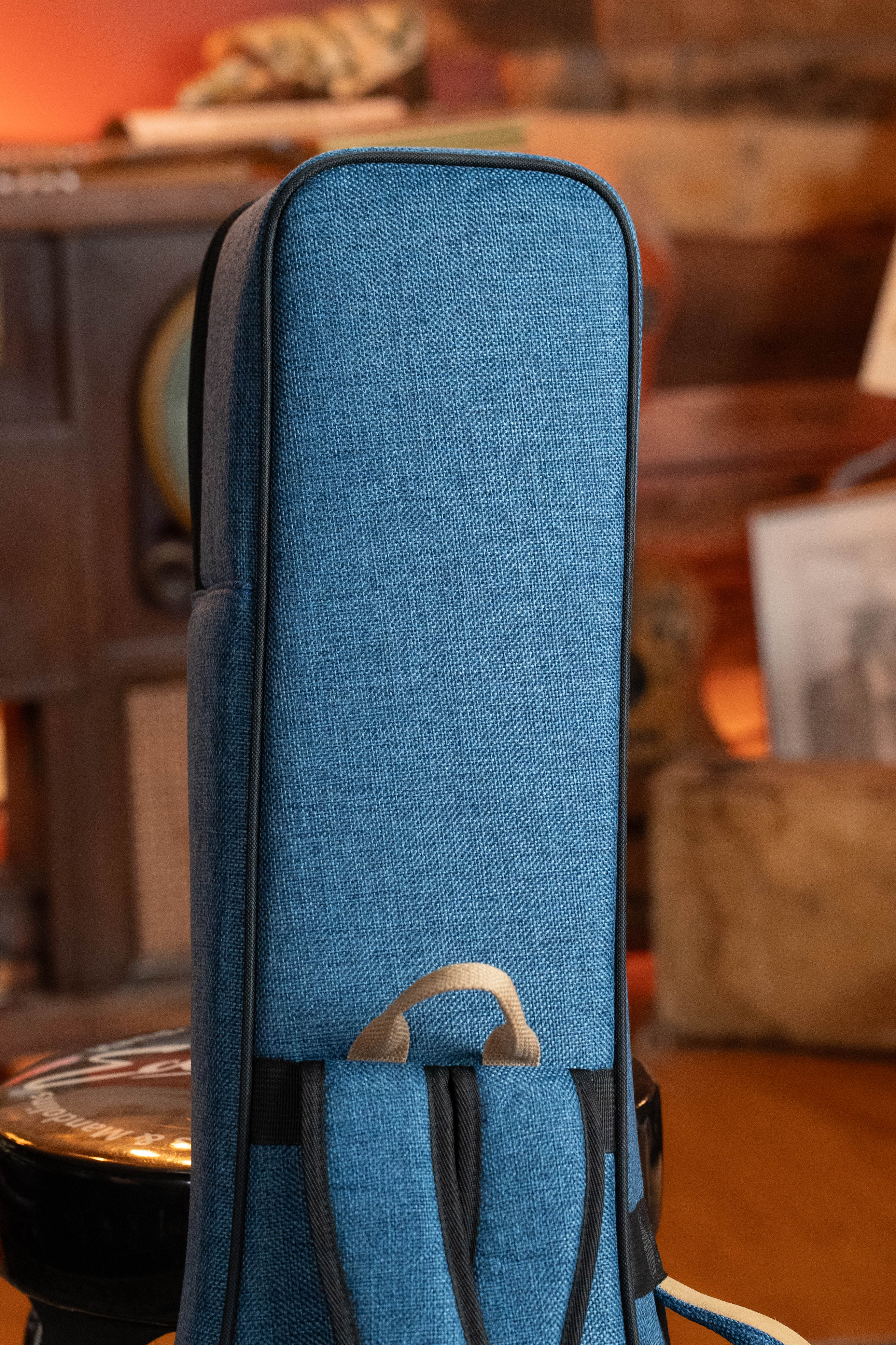A close-up of the Ortega OSOCABJ-OC Banjo Soft Case in ocean blue with a brown handle rests on a black stool, set against warmly lit wooden furniture and framed pictures.