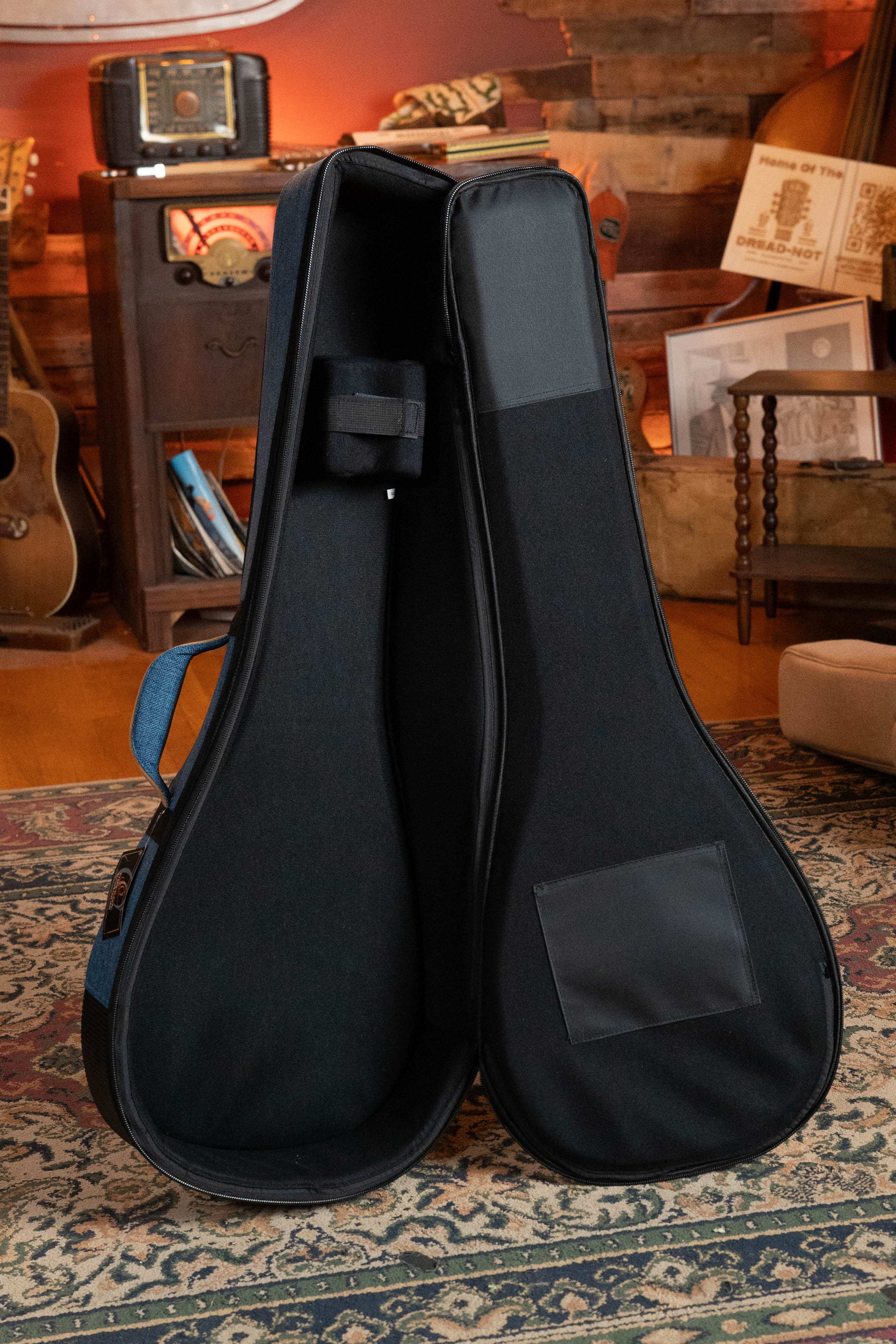An open black guitar case stands upright on a rug in a cozy room with guitars, wooden furniture, vintage decor, and an Ortega OSOCABJ-OC Banjo Soft Case in Ocean Blue by Ortega in the background.