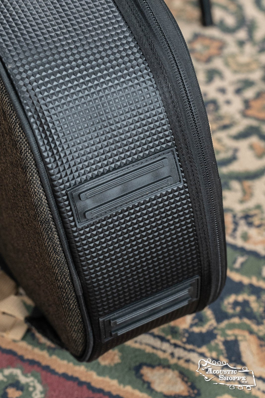 Close-up of the Ortega OSOCABJ-MO Banjo Soft Case in Mocca resting on a patterned rug, showing its textured surface, detailed ridges, and two rectangular plastic feet. The Acoustic Shoppe logo appears in the bottom right corner.