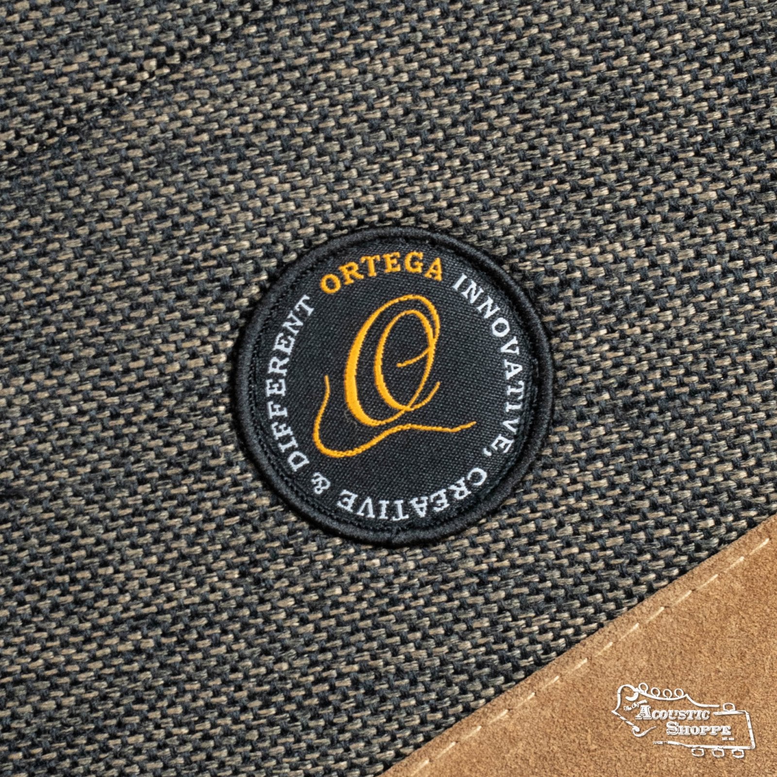 A black circular Ortega patch with orange and white text is sewn onto the textured gray suede OSOCADN-MO Dreadnought Guitar Soft Case in Mocca. The Acoustic Shoppe logo features in the bottom right corner.