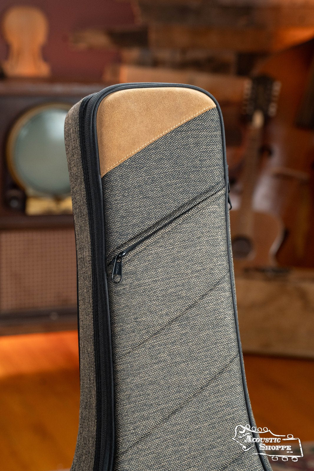 Close-up of the Ortega OSOCADN-MO Dreadnought Guitar Soft Case in Mocca, a gray padded gig bag with a brown suede accent and zippered pocket, shown indoors with blurred wooden furniture and instrument shapes.