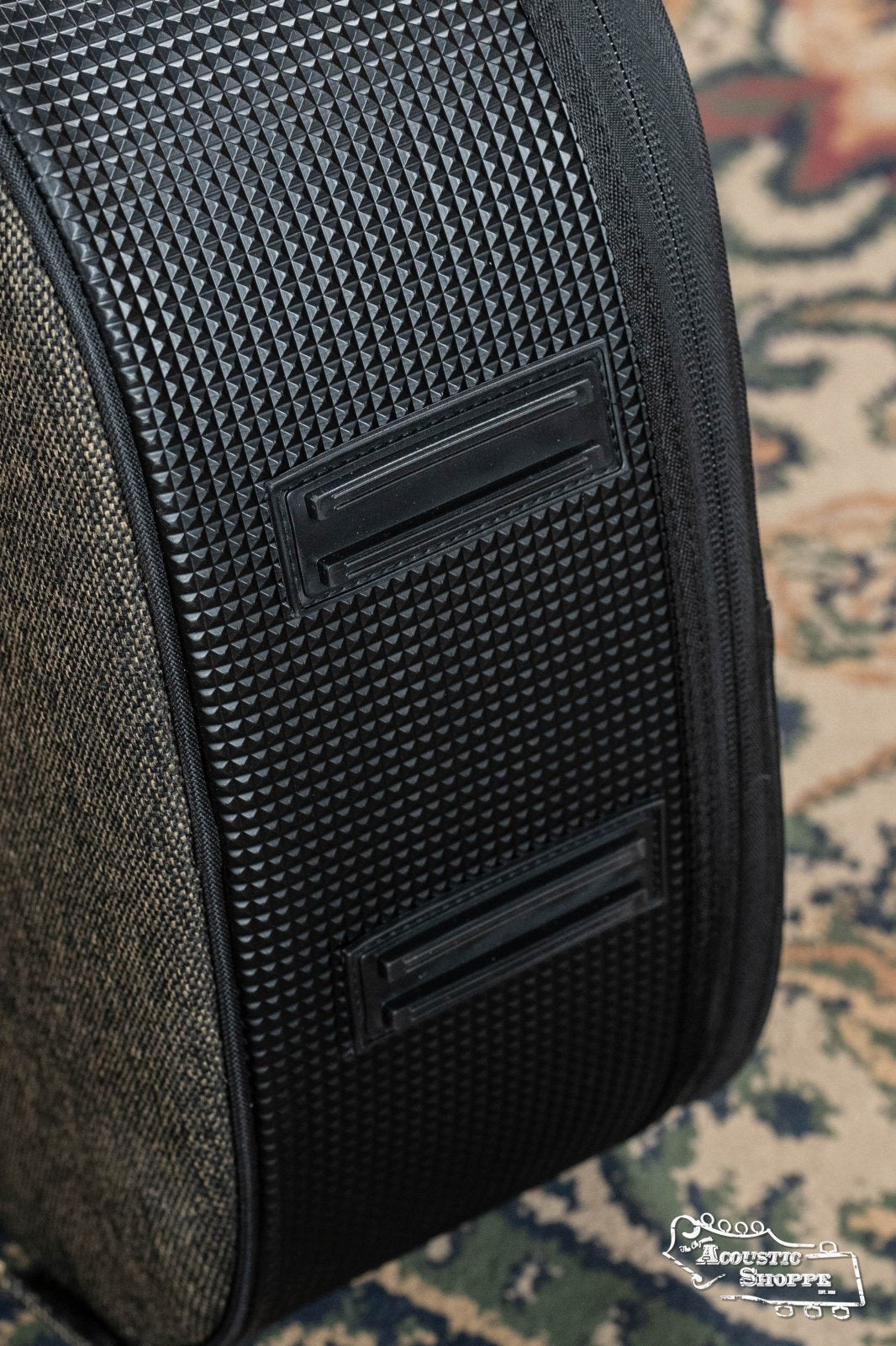 Close-up of the Ortega OSOCADN-MO Dreadnought Guitar Soft Case in Mocca, with two black handles, on a patterned rug. The bottom right features a white Acoustic Shoppe logo, contrasting with nearby padded gig bags. Brand: Ortega.
