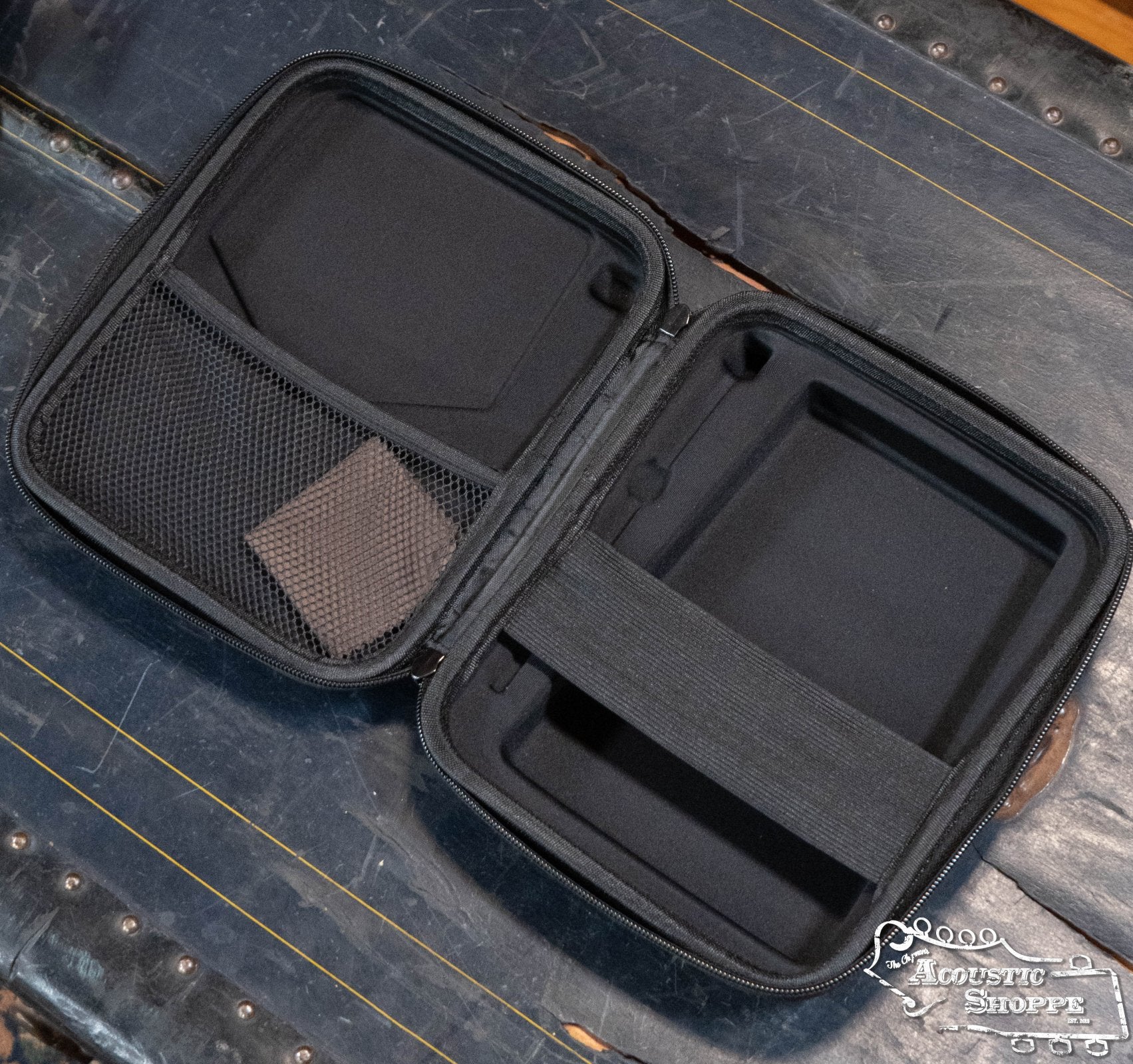 An open black hard-shell case with mesh pocket and elastic strap, shown empty on a blue wood surface. A small brown cleaning cloth is in the pocket—ideal for your Ortega OKB1E Lizard Series Solid Acacia Wood Kalimba w/ Built-In Pickup.