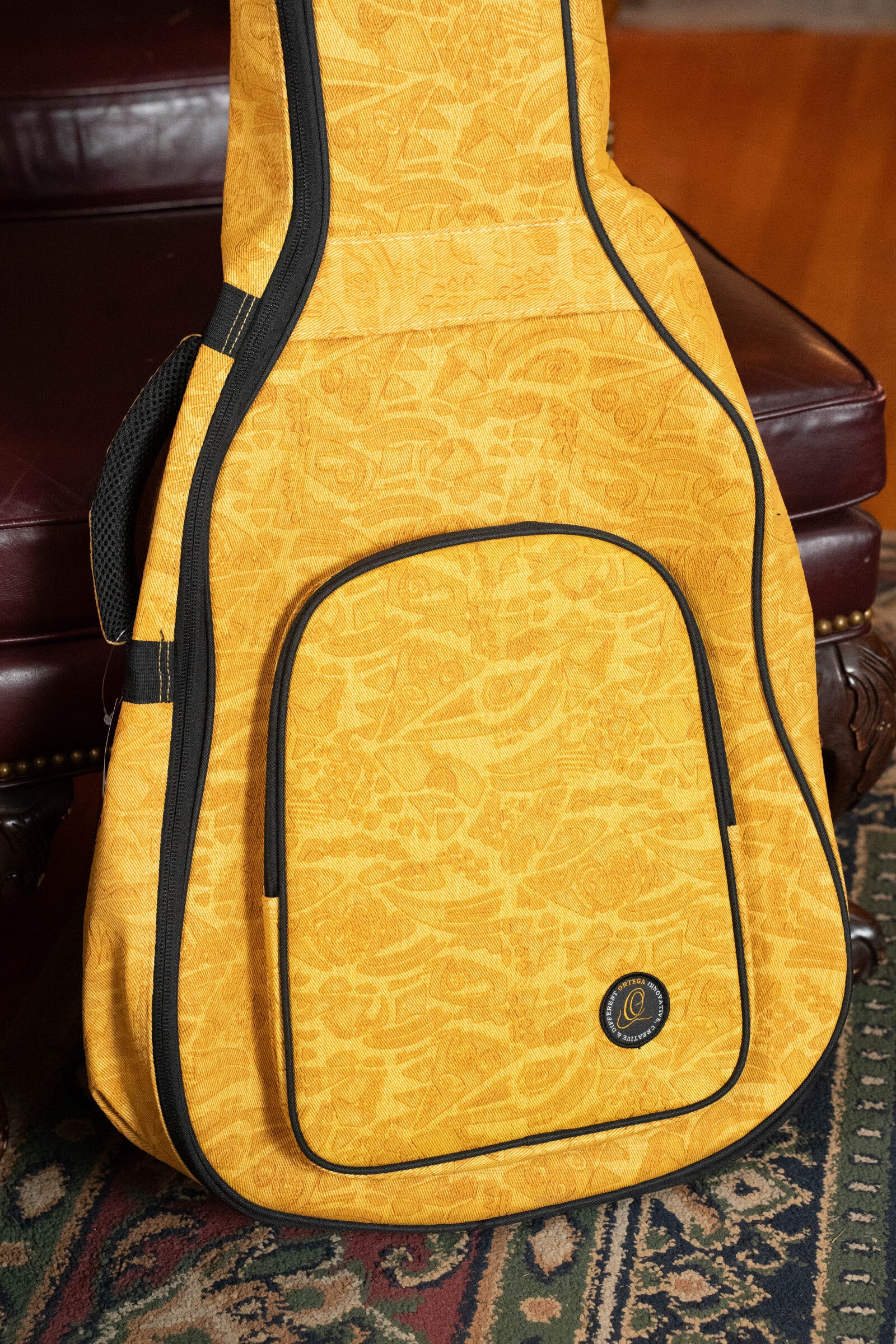 The Ortega OGBAC-DN-SUJ Dreadnought Guitar Gigbag in Sun Jean rests against a dark brown leather chair and a patterned rug.