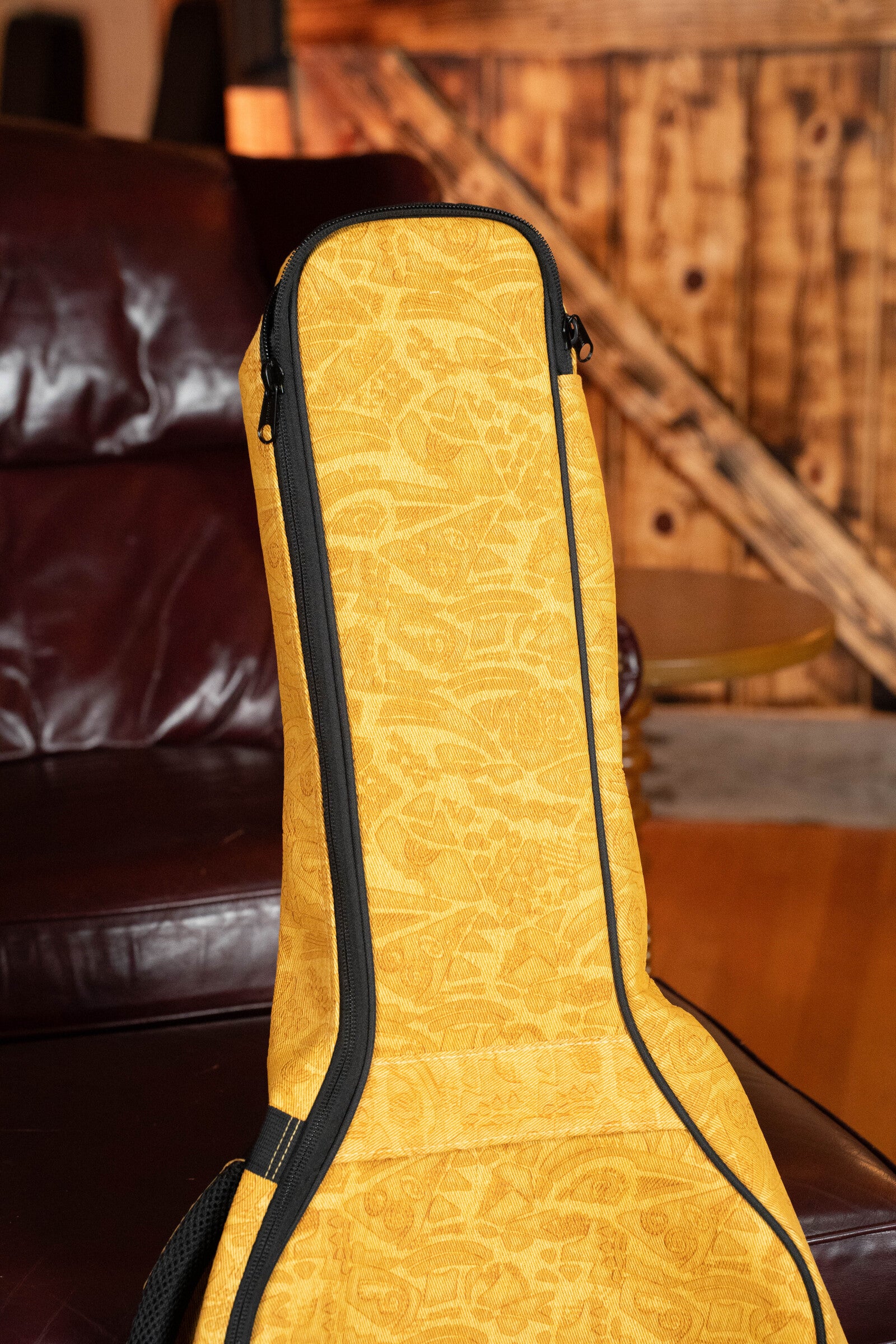 The Ortega OGBAC-DN-SUJ Dreadnought Guitar Gigbag in Sun Jean stands upright on a brown leather couch, featuring its subtle pattern, with a wooden wall and furniture visible in the background.