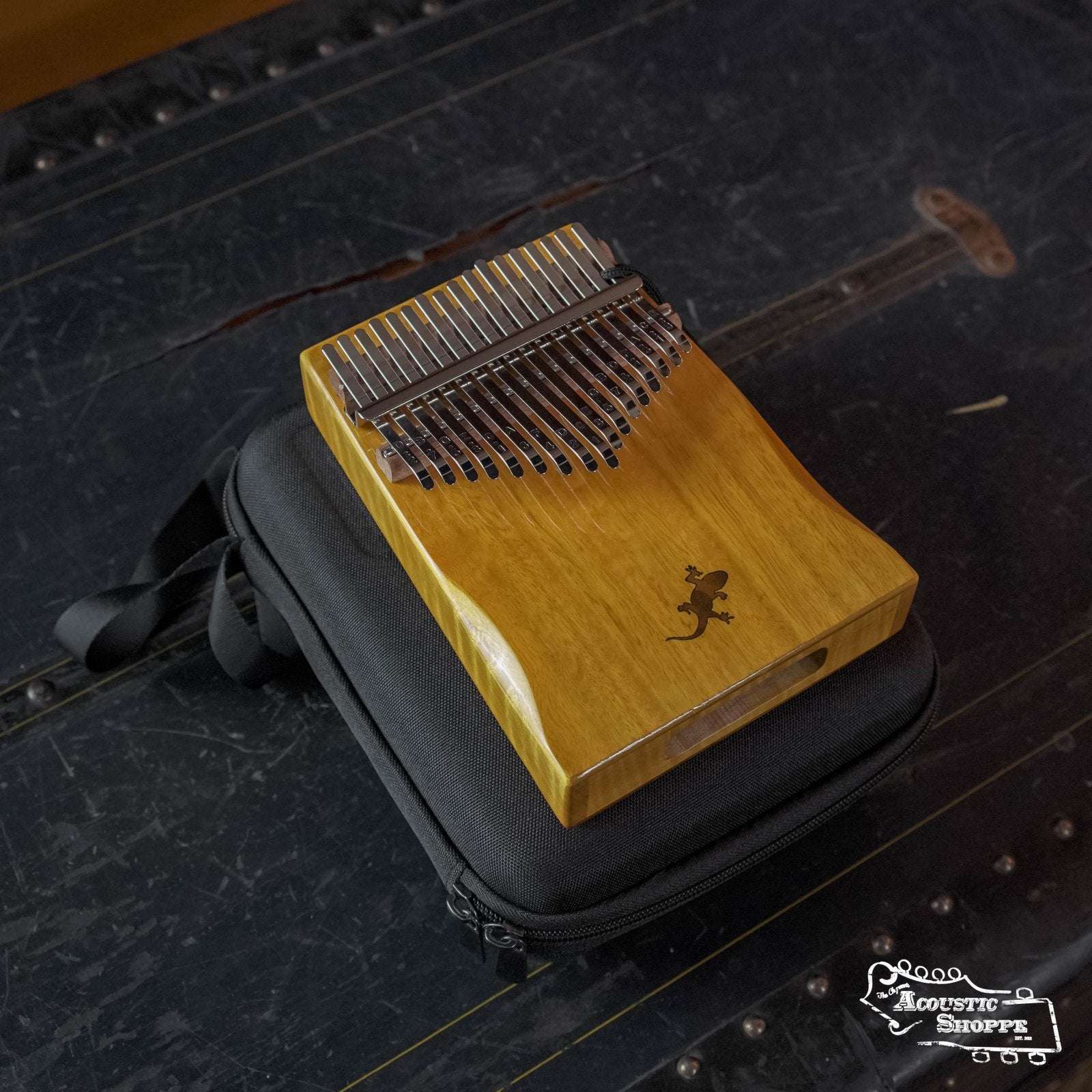 The Ortega OKB3 Lizard Series Solid Golden Phoebe Kalimba by Ortega, with 17 C Major keys and a lizard motif, is placed atop a black carrying case on a dark textured surface.