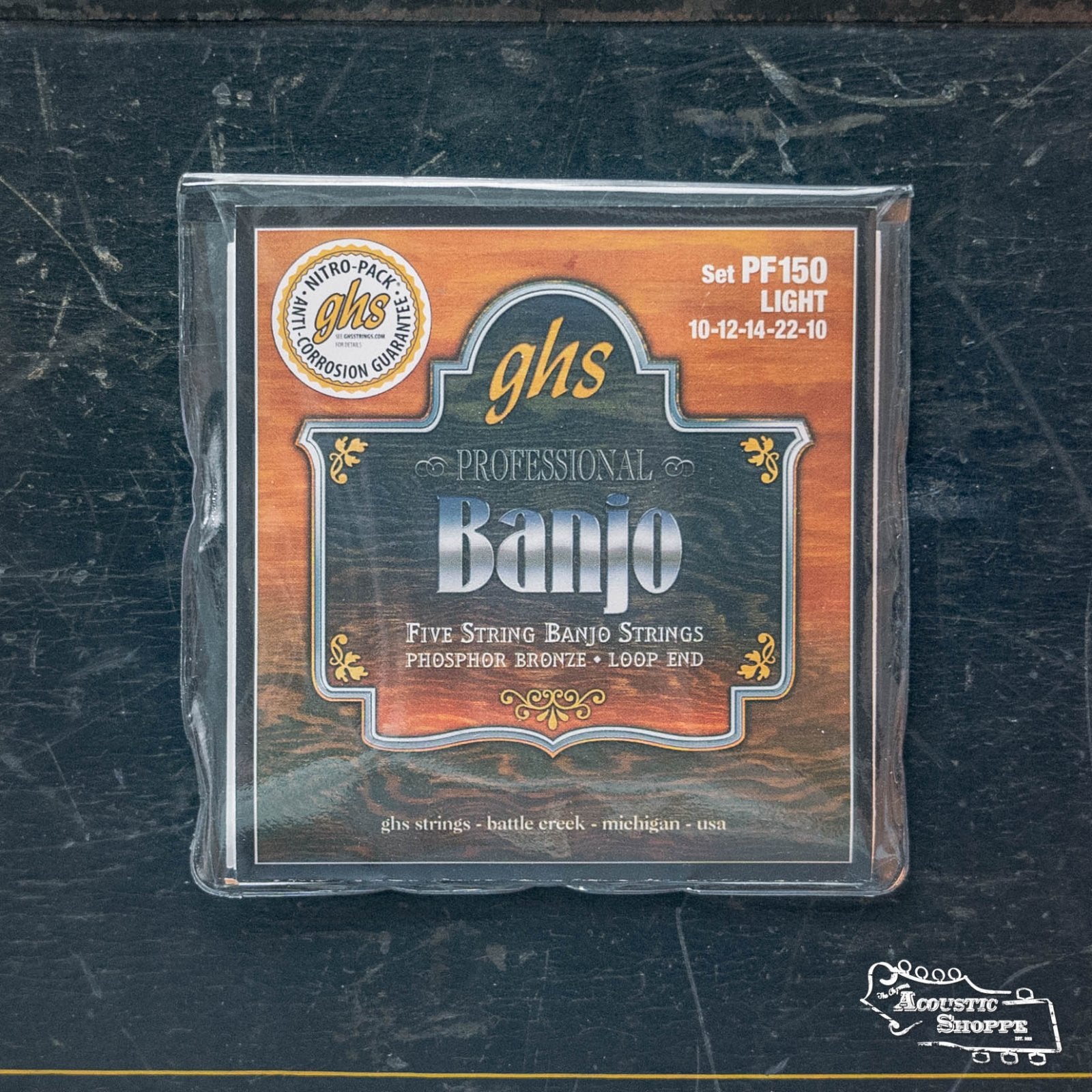 A package of GHS Strings PF150 Phosphor Bronze 5 String Banjo Strings - Light rests on a dark wooden surface, with The Acoustic Shoppe logo visible in the lower right corner.