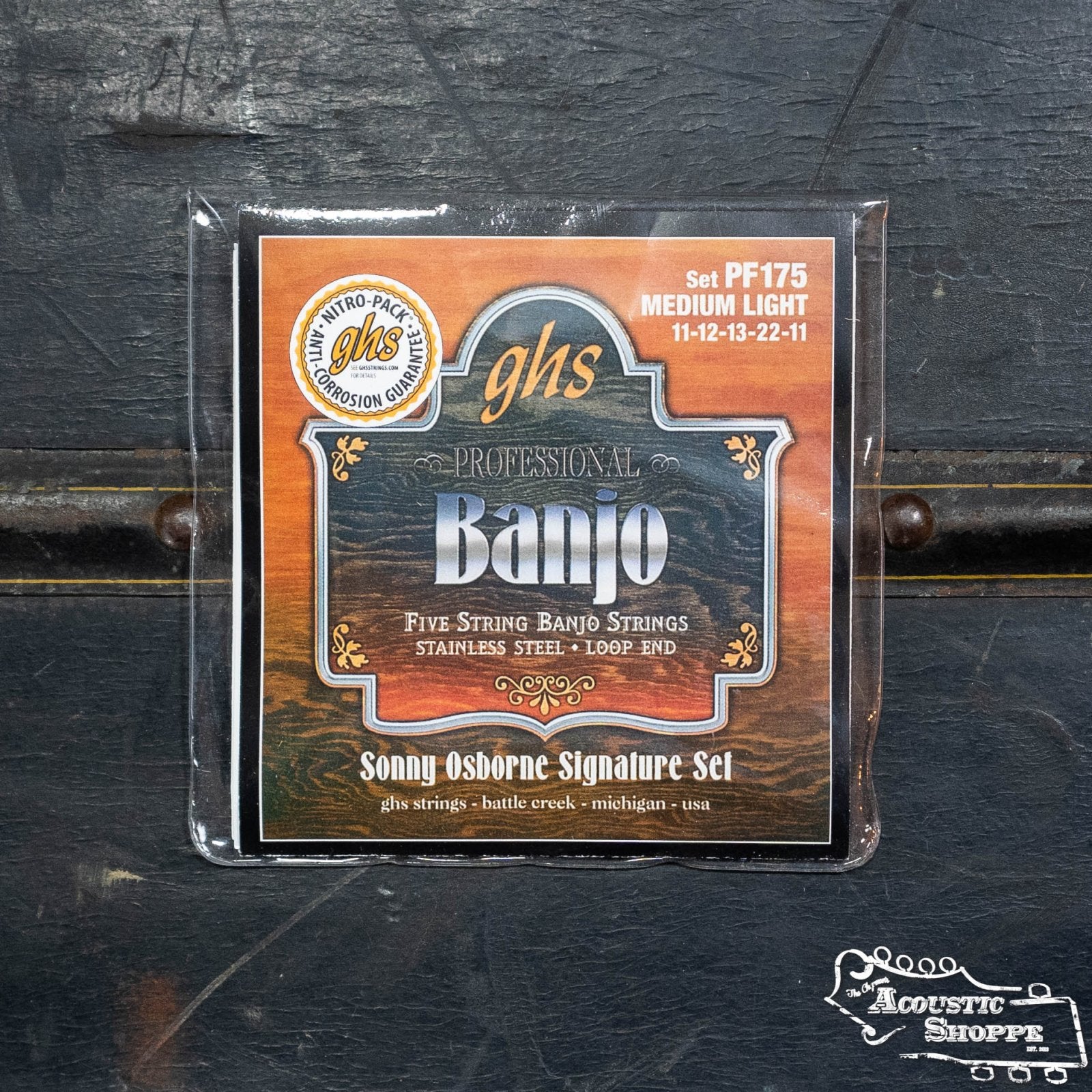A pack of GHS Strings PF175 Sonny Osborne Signature Stainless Steel 5-String Banjo Strings (Regular Light) lies on a dark, textured surface. This set includes gauges 11-12-13-22-11 with plain steel banjo strings.