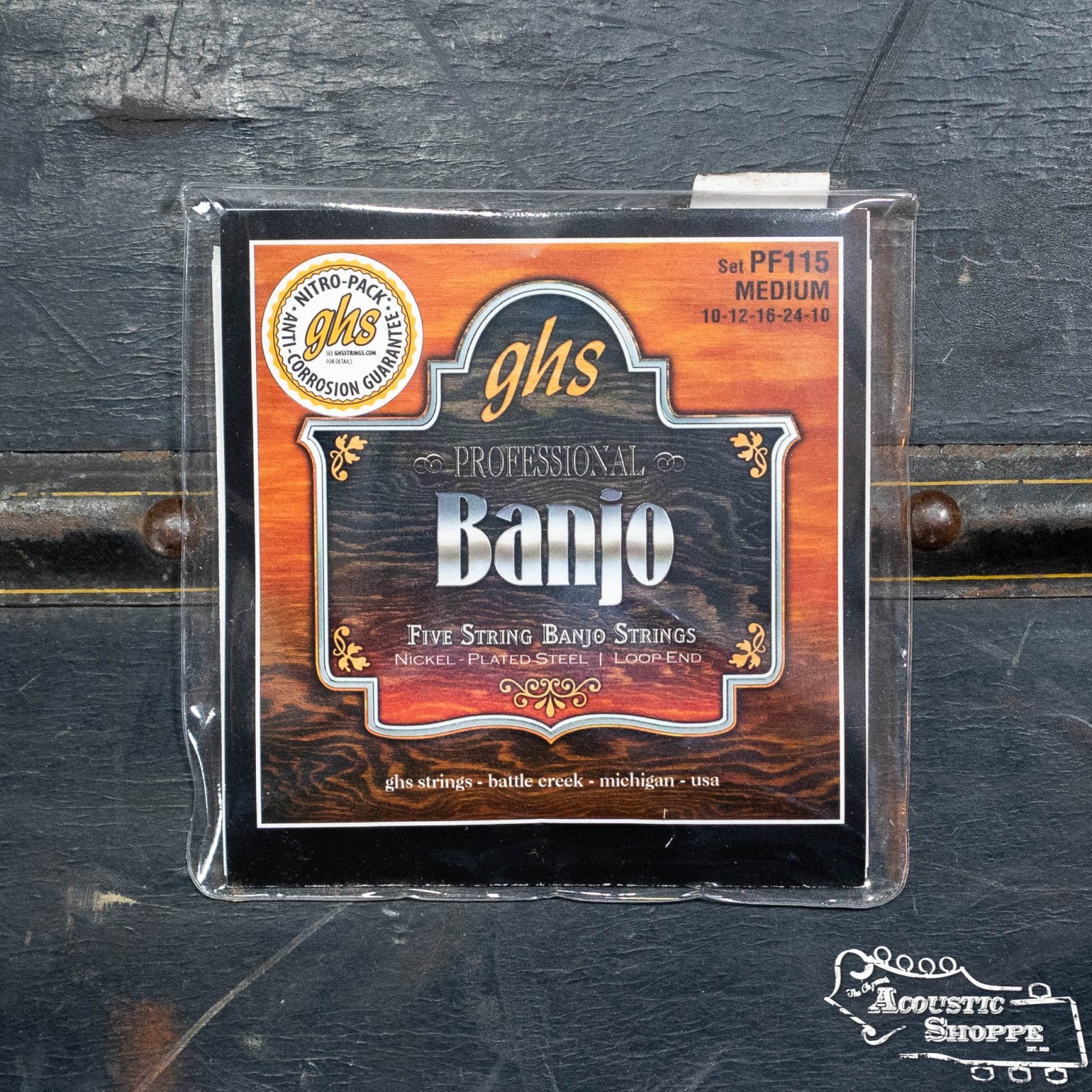 A pack of GHS Strings PF115 5 String Banjo Nickel Plate Medium (gauges 10-12-16-24-10) is shown on a dark wooden surface, with “The Acoustic Shoppe” logo in the corner.