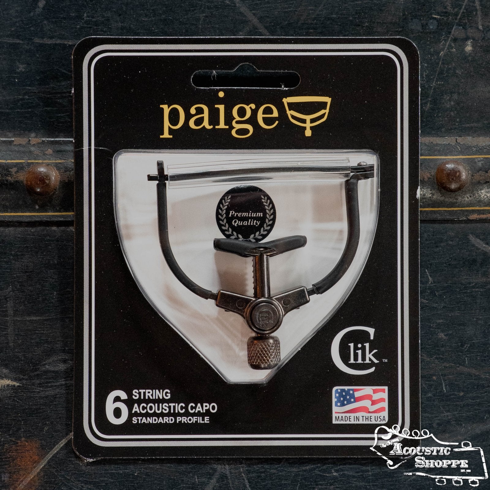 The Paige Clik 6-String Guitar Capo (PC-6-2.062-R) by Paige Musical Products is a standard profile, premium quality capo for acoustic guitars. Made in the USA, it comes in packaging with The Acoustic Shoppe logo at the lower right corner.