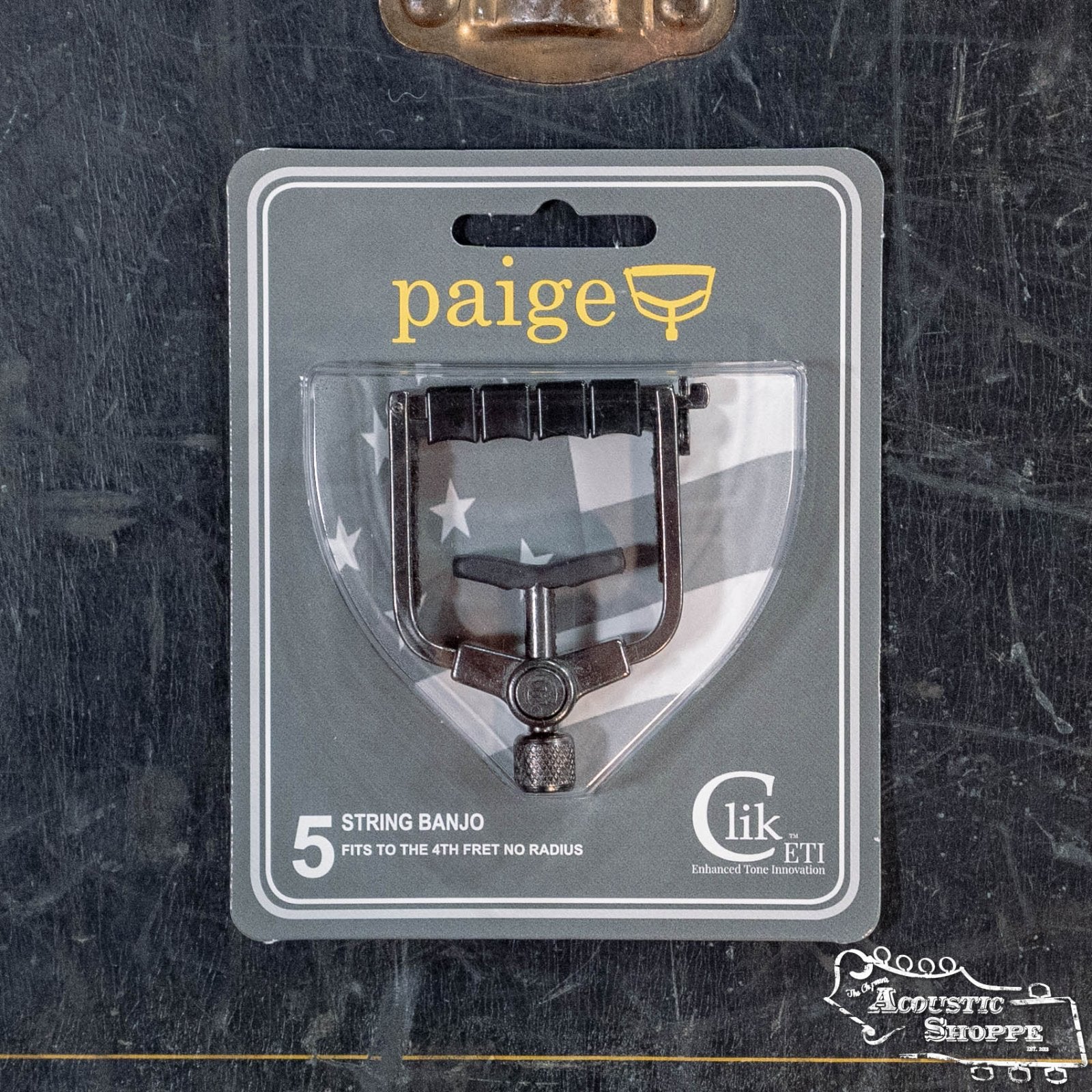 The Paige Musical Products Click ETI 5-String Banjo Capo (PC-B4-ETI), designed for 4th fret max/no radius, comes in gray and clear packaging highlighting enhanced tone innovation and is available from The Acoustic Shoppe.