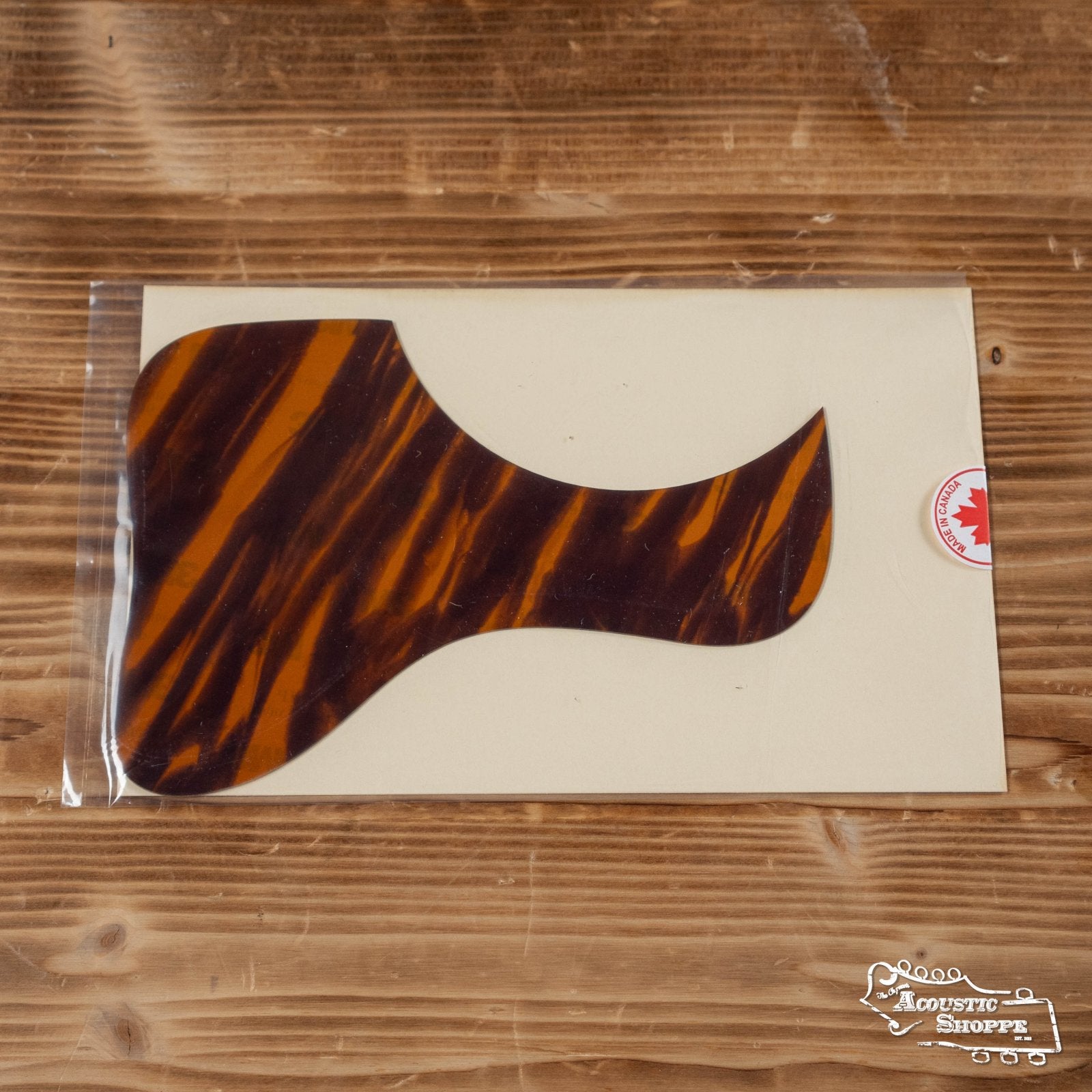 The Proulx Lutherie Tortoloid AFS-LOO Hand-Poured Custom Amber Firestripe L-OO Style Pickguard, featuring a curved design, is sealed in clear plastic packaging and rests on a wooden surface.