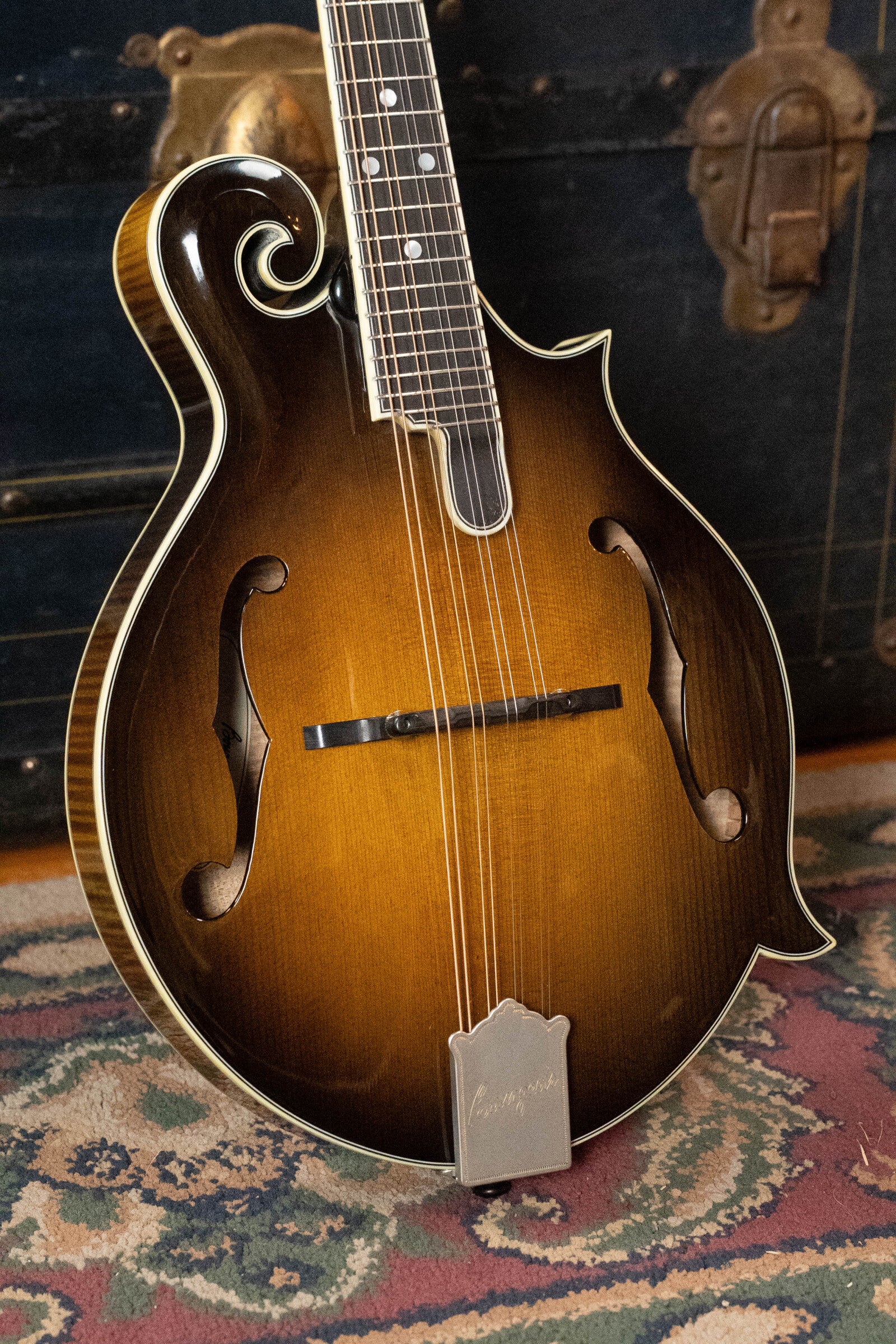 A Bourgeois *Pre-Order 25% Deposit ONLY* M5-F-SB/M5-A-SB Aged-Tone Adirondack/Maple Sunburst Mandolin stands upright on a patterned rug before a vintage trunk with brass accents.