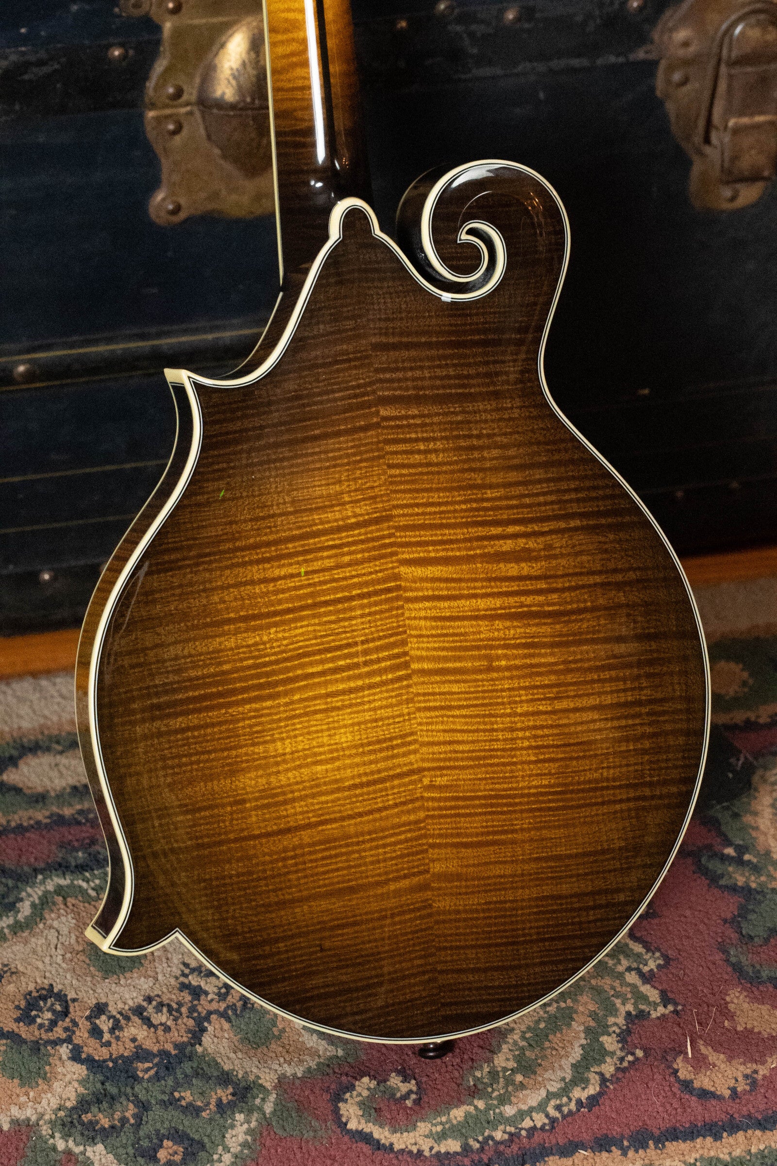 A close-up of the back of a Bourgeois M5-F-SB Aged-Tone Adirondack/Maple Sunburst F-Style Mandolin with decorative white binding, resting on a patterned rug in front of a vintage trunk. *Pre-Order 25% Deposit ONLY*.