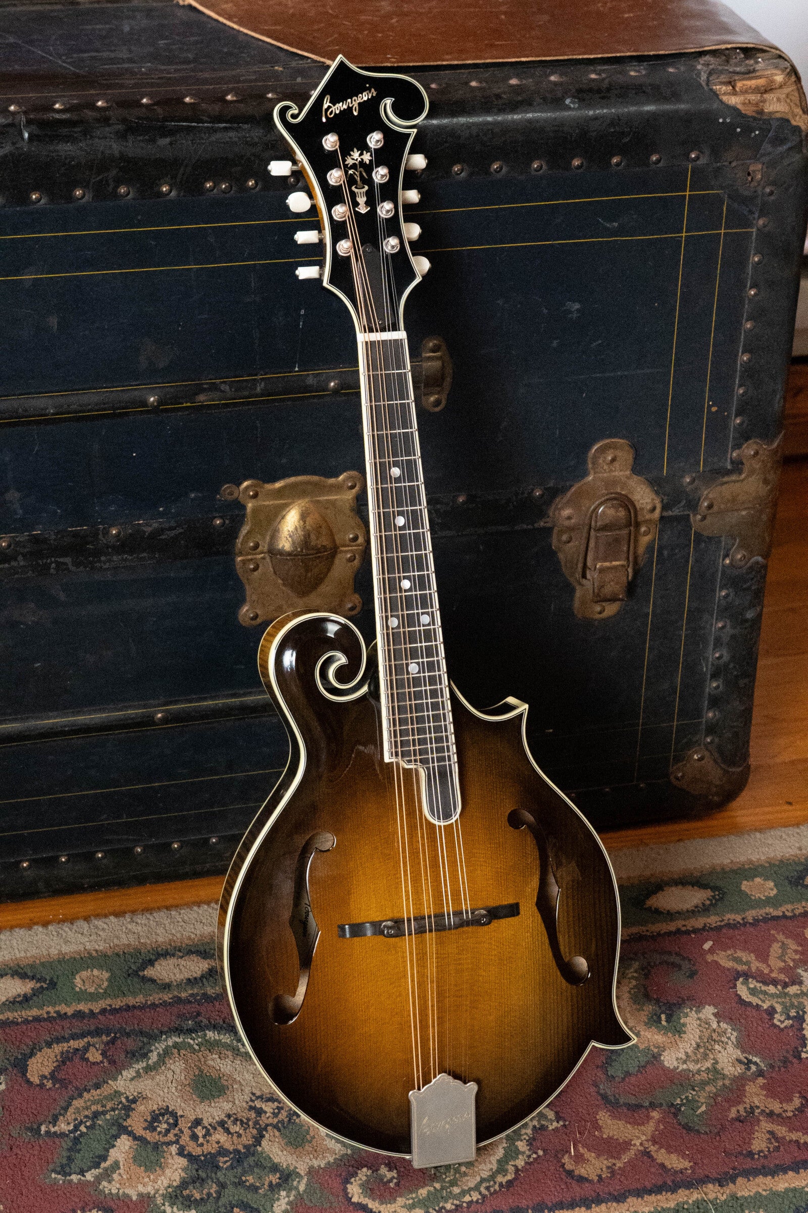 A Bourgeois *Pre-Order 25% Deposit ONLY* M5-F-SB/M5-A-SB Aged-Tone Adirondack/Maple Sunburst Mandolin leans by a vintage black trunk with brass hardware, set on a patterned rug over a wooden floor.