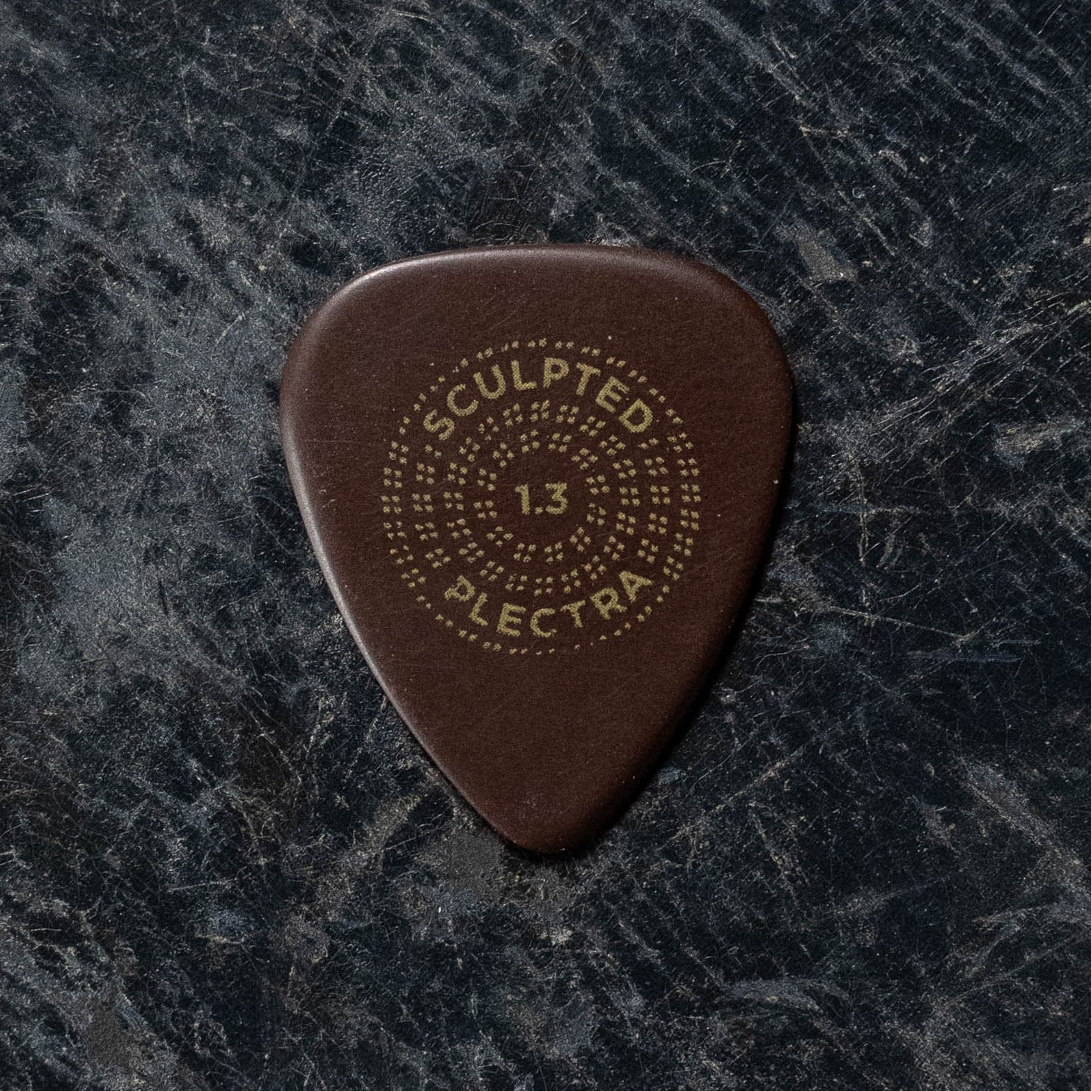 A Dunlop Primetone Standard Smooth Pick from the 3 Pack (511P) lies on a scratched, dark surface, its beveled brown edge and gold text echoing the classic style of this trusted Dunlop guitar accessory.