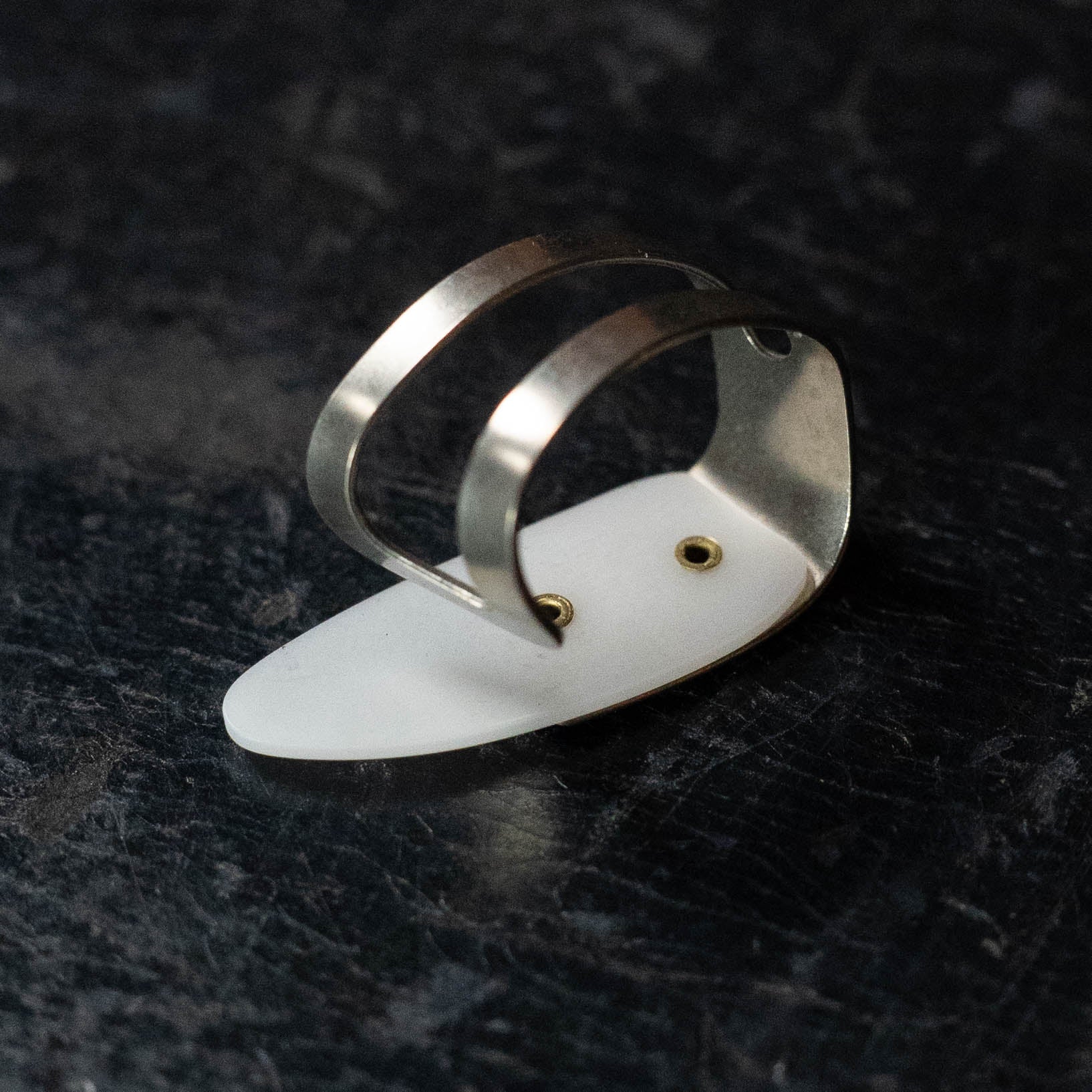 A close-up of the ProPik Metal/Plastic Thumbpick Small (11-206-3004) by ProPik, featuring a modern design with two parallel silver bands and a white oval base, resting on a dark textured surface.