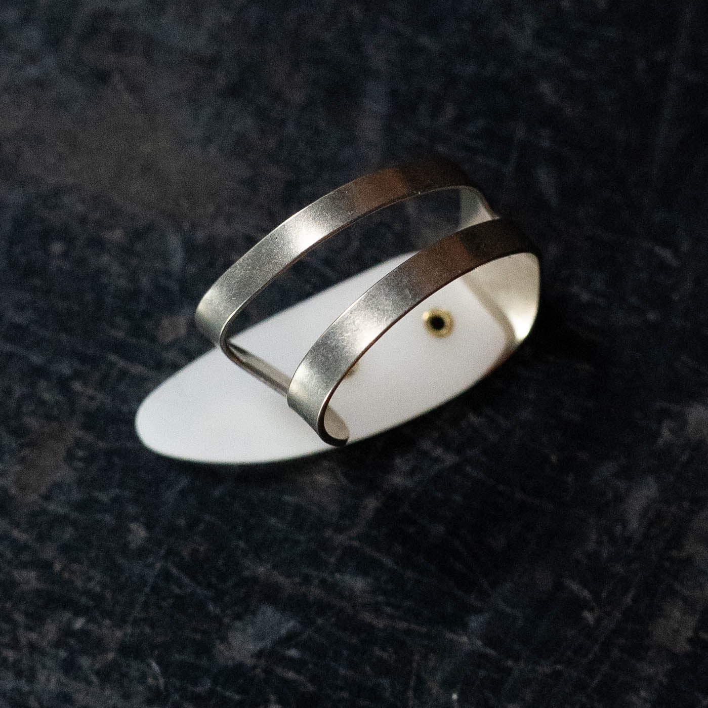 The ProPik Metal/Plastic Thumbpick Small (11-206-3004) by ProPik is displayed on a white oval stand, set against a dark, textured background. Its modern design features sleek, parallel bands reminiscent of fingerpicks.