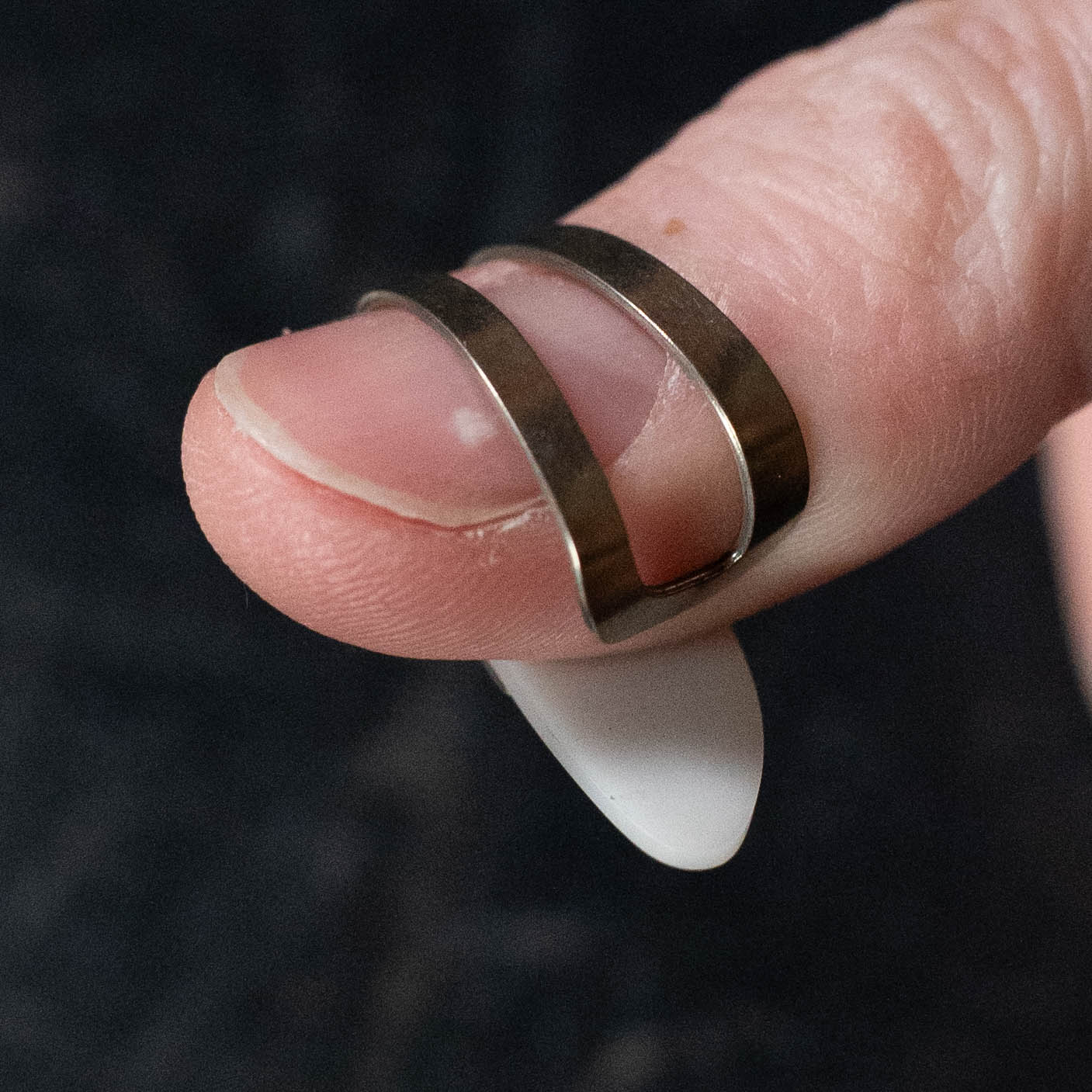 A close-up of a hand wearing the ProPik Metal/Plastic Thumbpick Small (11-206-3004) by ProPik on the thumb, with a white almond-shaped artificial nail, set against a dark, blurred background.