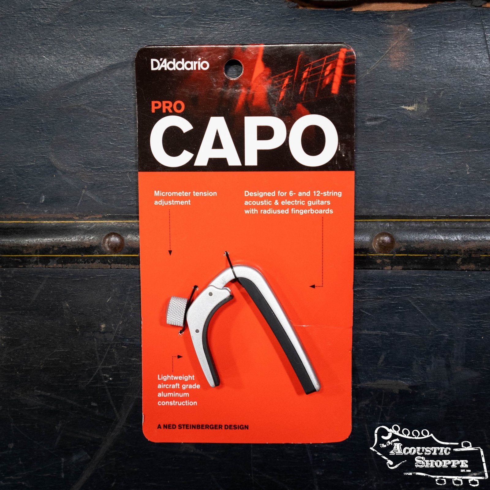 The D'Addario NS Capo Pro PW-CP-02 is a silver guitar capo with a black grip, packaged to highlight its micrometer tension adjustment and lightweight aluminum build.