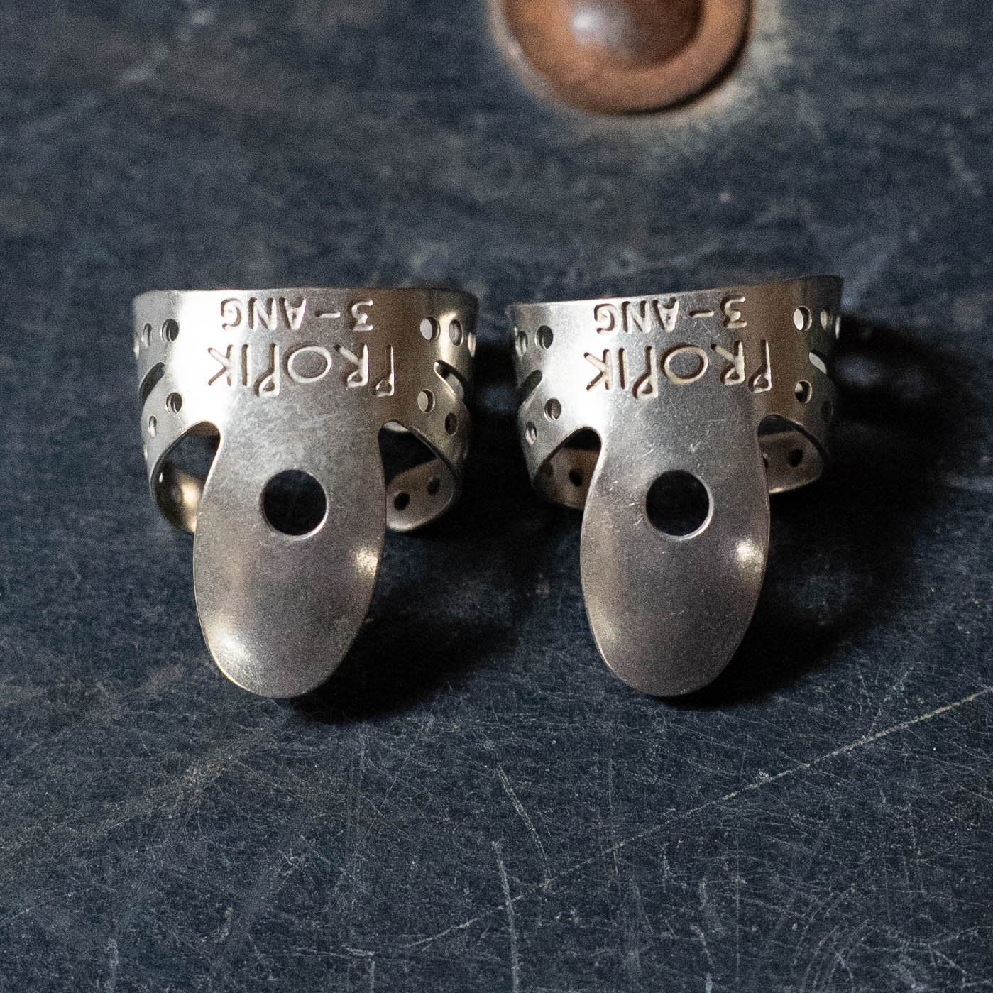 Two ProPik Angled Extra Comfortable Nickel Split Wrap #3 Fingerpicks (2-Pack, 11-201-1432) by ProPik rest on a textured dark surface. The picks have holes and an oval-shaped tip extension.