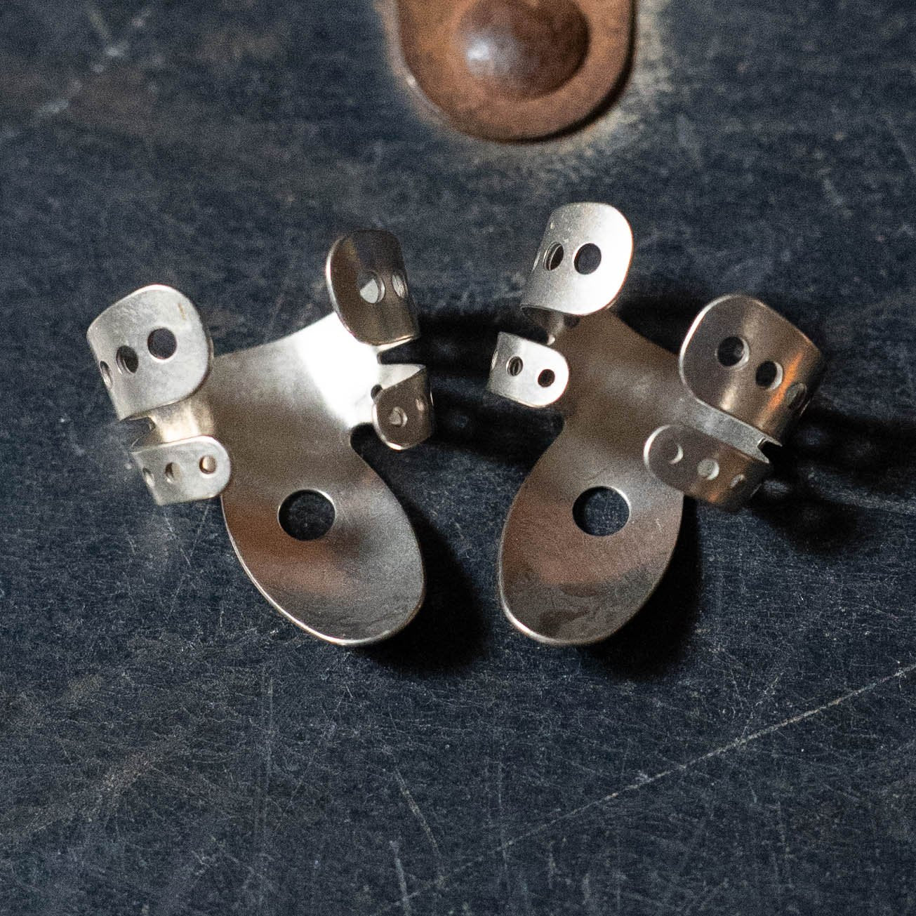 Two ProPik Angled Extra Comfortable Nickel Split Wrap #3 Fingerpicks (2-pack) by ProPik, featuring multiple cut-out holes and a unique wrap design, rest on a dark textured surface with a small round brown detail above.