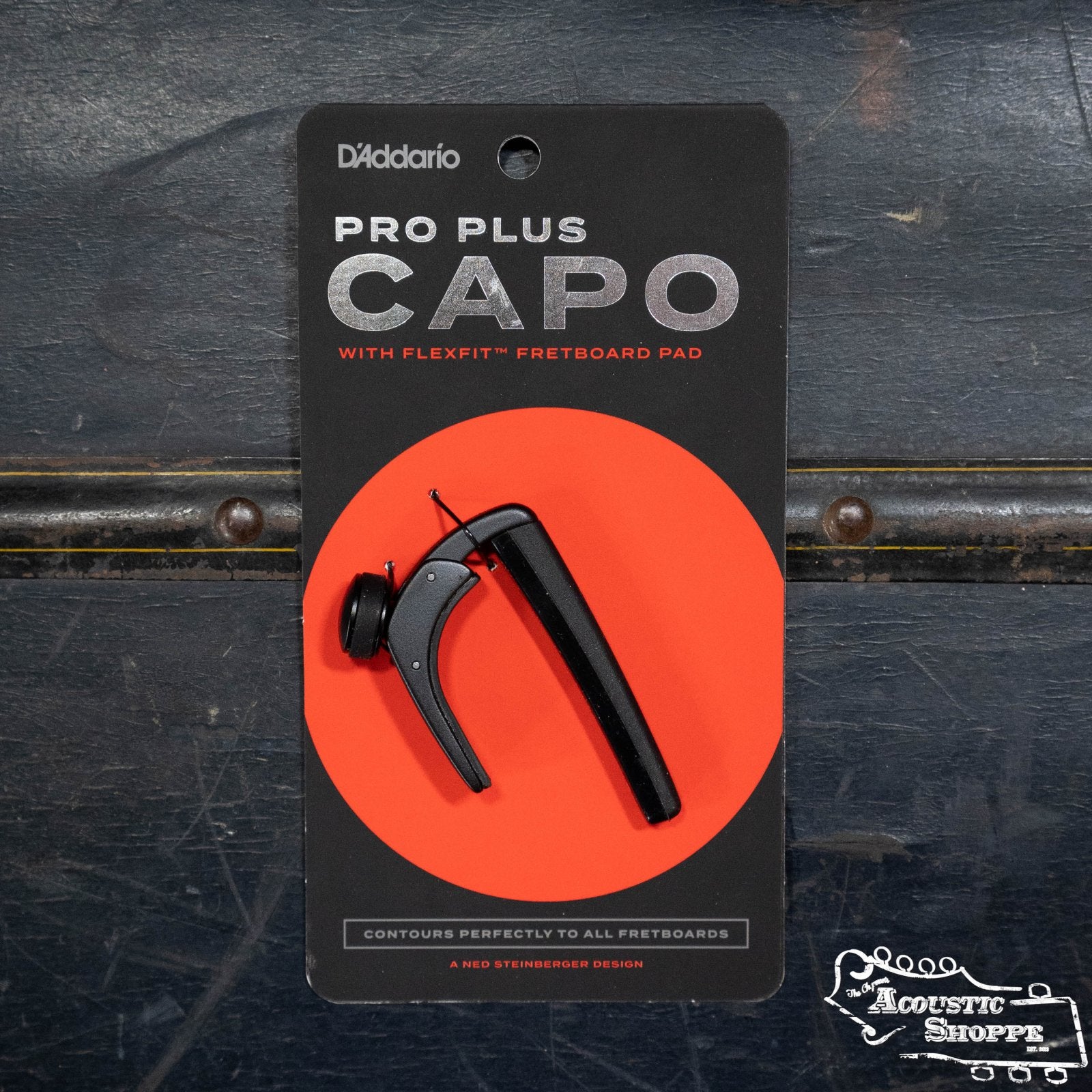 A D'Addario Pro Plus Guitar Capo in black is shown in branded packaging with an orange circle background on a dark textured surface. The packaging includes product details and The Acoustic Shoppe logo.