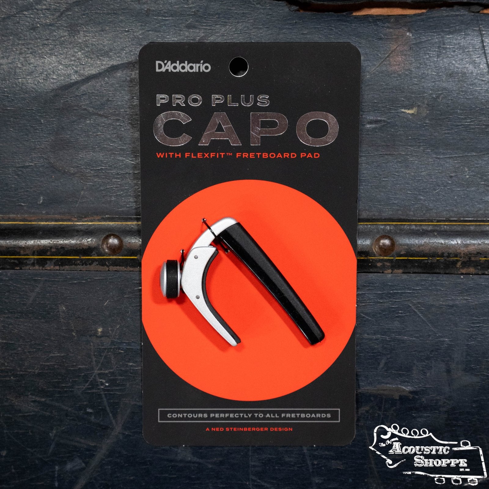 A D'Addario Pro Plus Guitar Capo - Silver with FlexFit technology, in black and orange packaging featuring white and orange branding, rests on a dark textured surface.