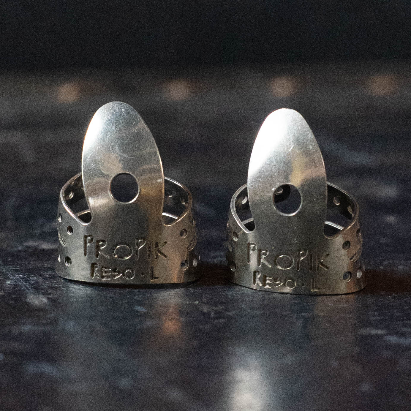 Two ProPik Reso Nickel Split Wrap Large Finger Picks (2-Pack, 11-203-1412) stand upright on a dark, textured surface, displaying their circular holes and curved tips—perfect for resophonic guitar players.