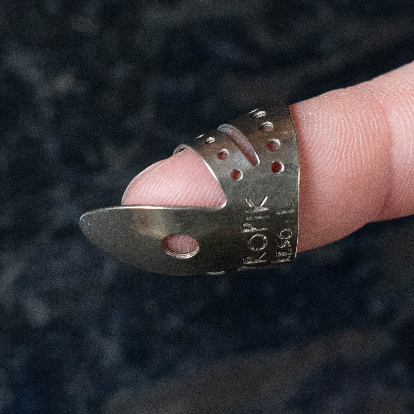 A ProPik Reso Nickel Split Wrap Large Finger Pick (11-203-1412) by ProPik, worn on a person's fingertip, is shown against a dark background. This metal pick features a curved, pointed end and comes in a 2-pack.
