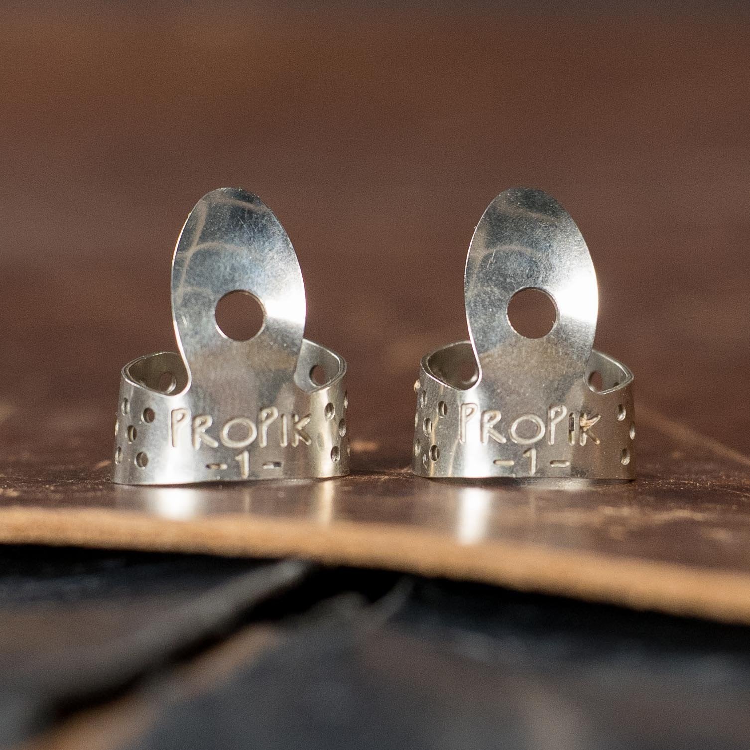 Two ProPik Standard Nickel Single Wrap #1 Finger Picks (2-Pack) by Deering Banjos stand upright on leather, showing engraved text and holes on each adjustable pick.