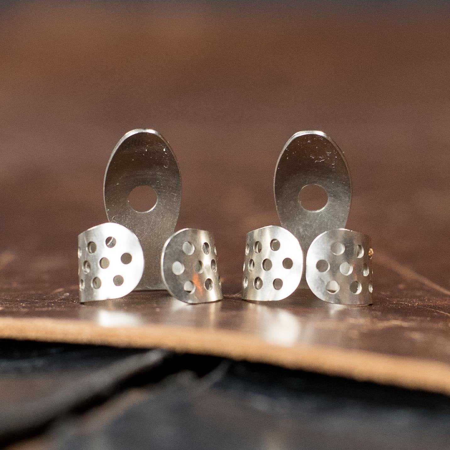 A set of ProPik Standard Nickel Single Wrap #1 Finger Picks (2-Pack) by Deering Banjos stands upright on a leather surface, their abstract, perforated design reminiscent of vintage silver earrings with circular cutouts.