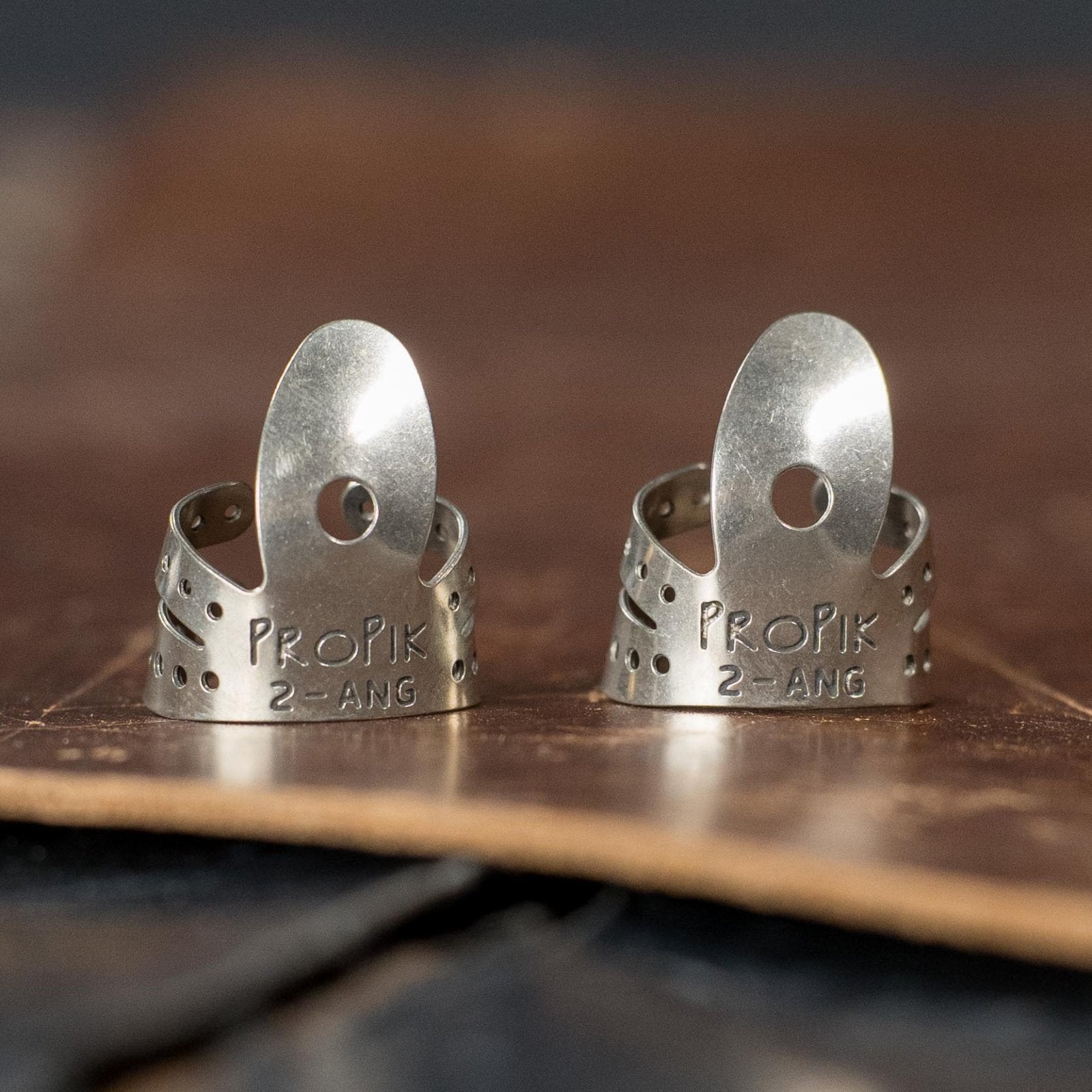 A (2-Pack) of ProPik Angled Extra Comfortable Nickel Split Wrap #2 Fingerpicks (11-201-1422) from ProPik, featuring adjustable perforated sides and a curved tip, rests on leather—ideal for banjo or guitar players.