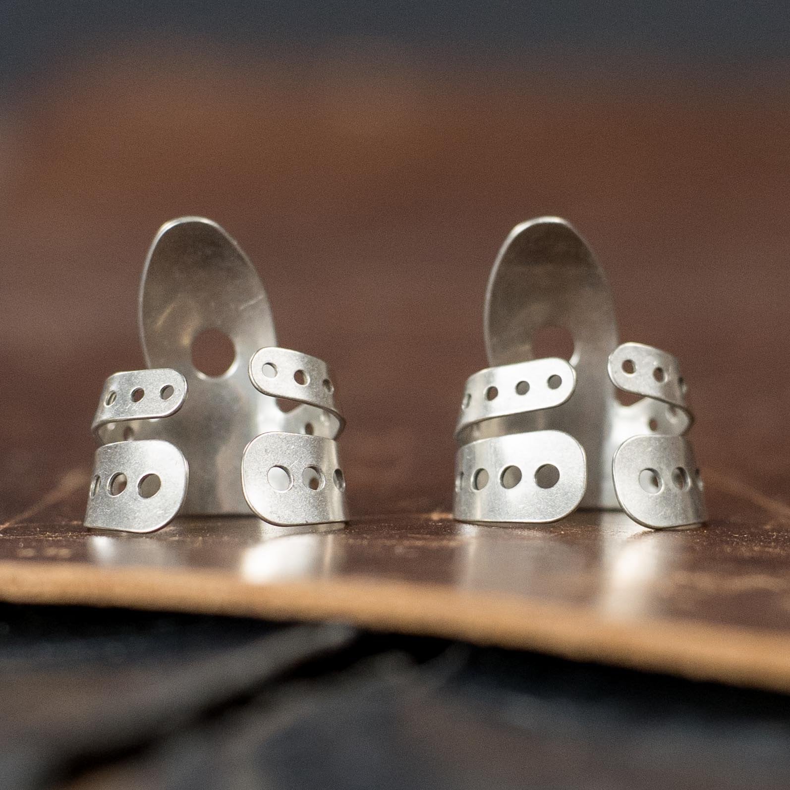 A pair of ProPik Angled Extra Comfortable Nickel Split Wrap #1 Finger Picks (2-pack) by ProPik, featuring a layered, adjustable design with multiple holes, shown upright on brown leather for a modern, industrial look.