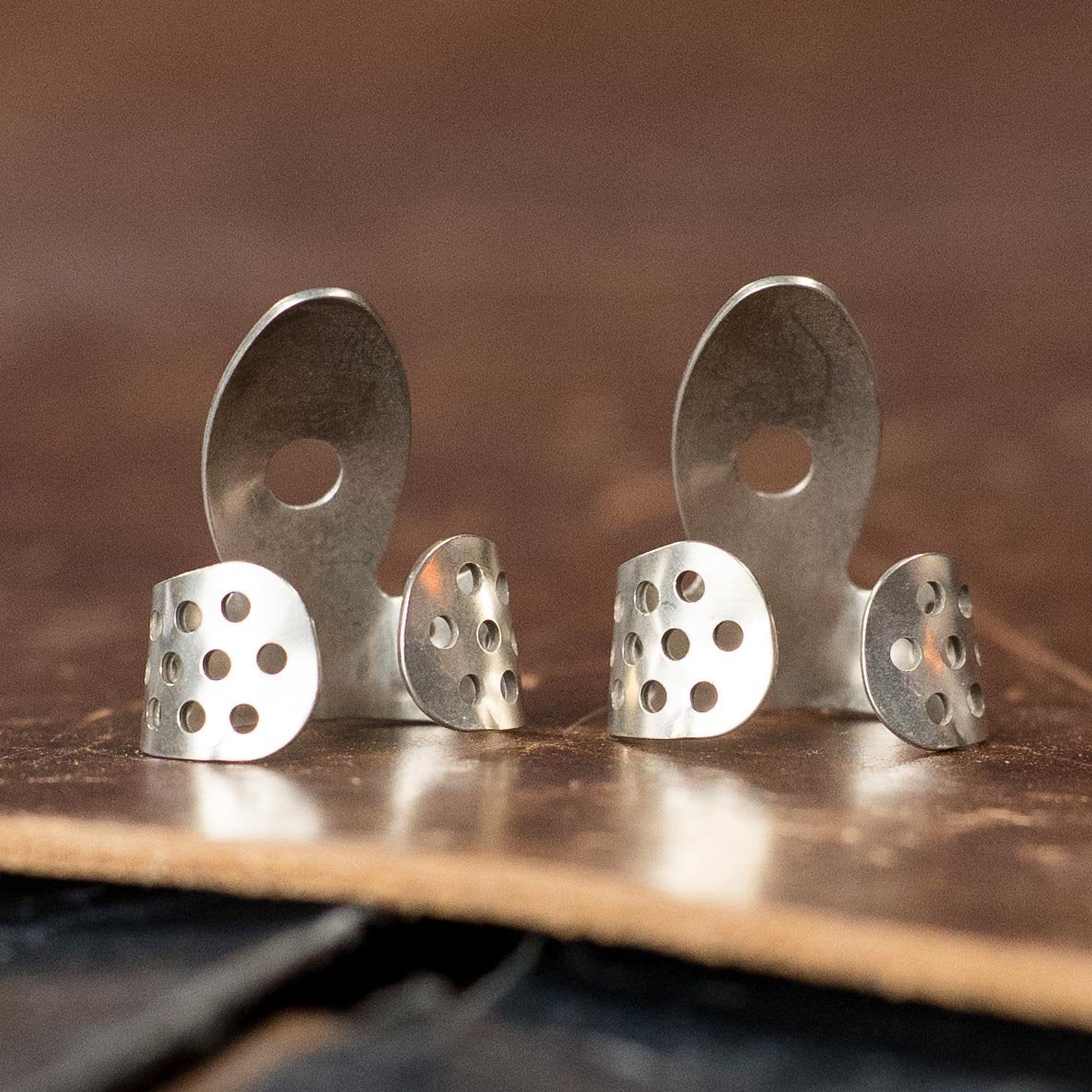 A (2-Pack) of ProPik Nickel Single Wrap Medium Finger Picks (11-202-1113) by ProPik is shown, featuring a modern, sleek design with circular and oval cut-out shapes, displayed on a wooden surface.