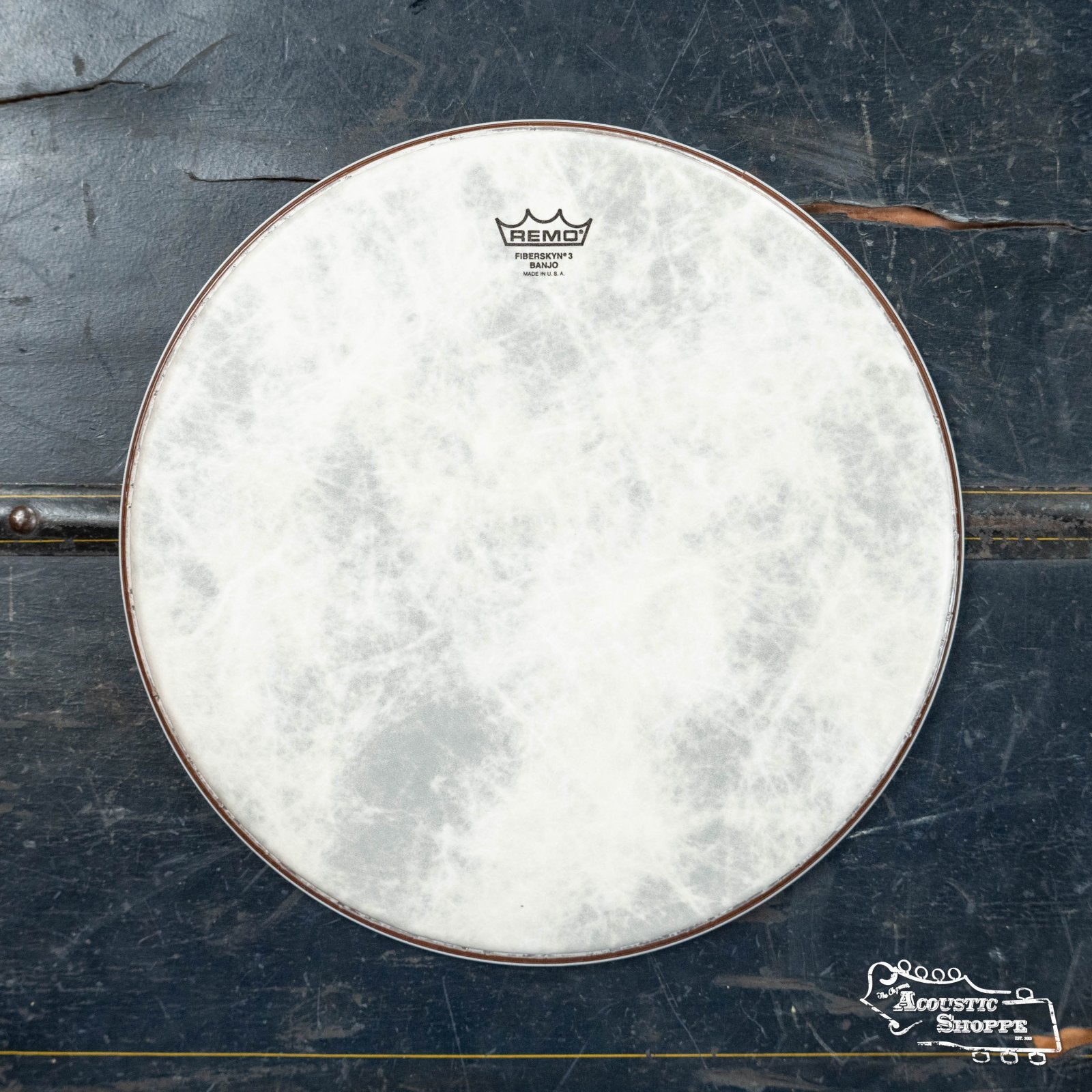 A Remo Banjo Fiberskyn Medium Collar 10-11/16in drumhead with a textured white surface is centered on worn dark wood; the Remo logo appears at the top, with a small shop logo in the lower right corner.