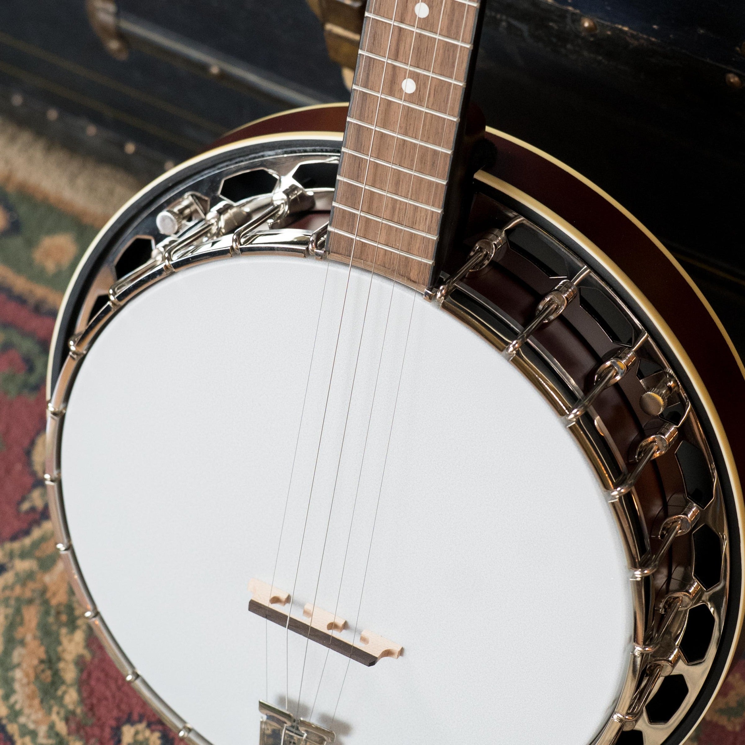 Close-up of a Recording King RKH-05 Dirty 30s Resonator Banjo #2563 upright, displaying its white drum head, strings, dot-inlay wooden neck, and metal hardware; a patterned rug and dark wooden trunk appear in the background.