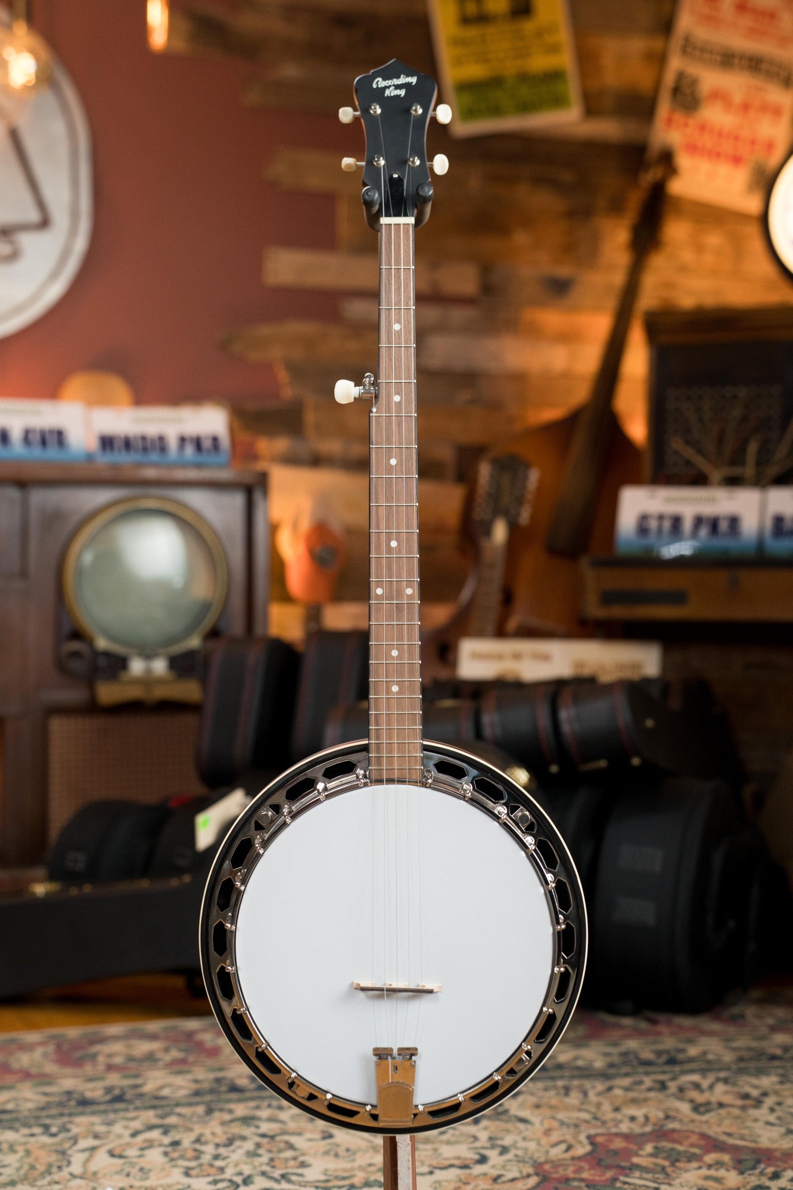 A Recording King RKH-05 Dirty 30s Resonator Banjo #2563 by Recording King stands upright on a stand in a warmly lit room surrounded by musical instruments, cameras, and vintage signs.