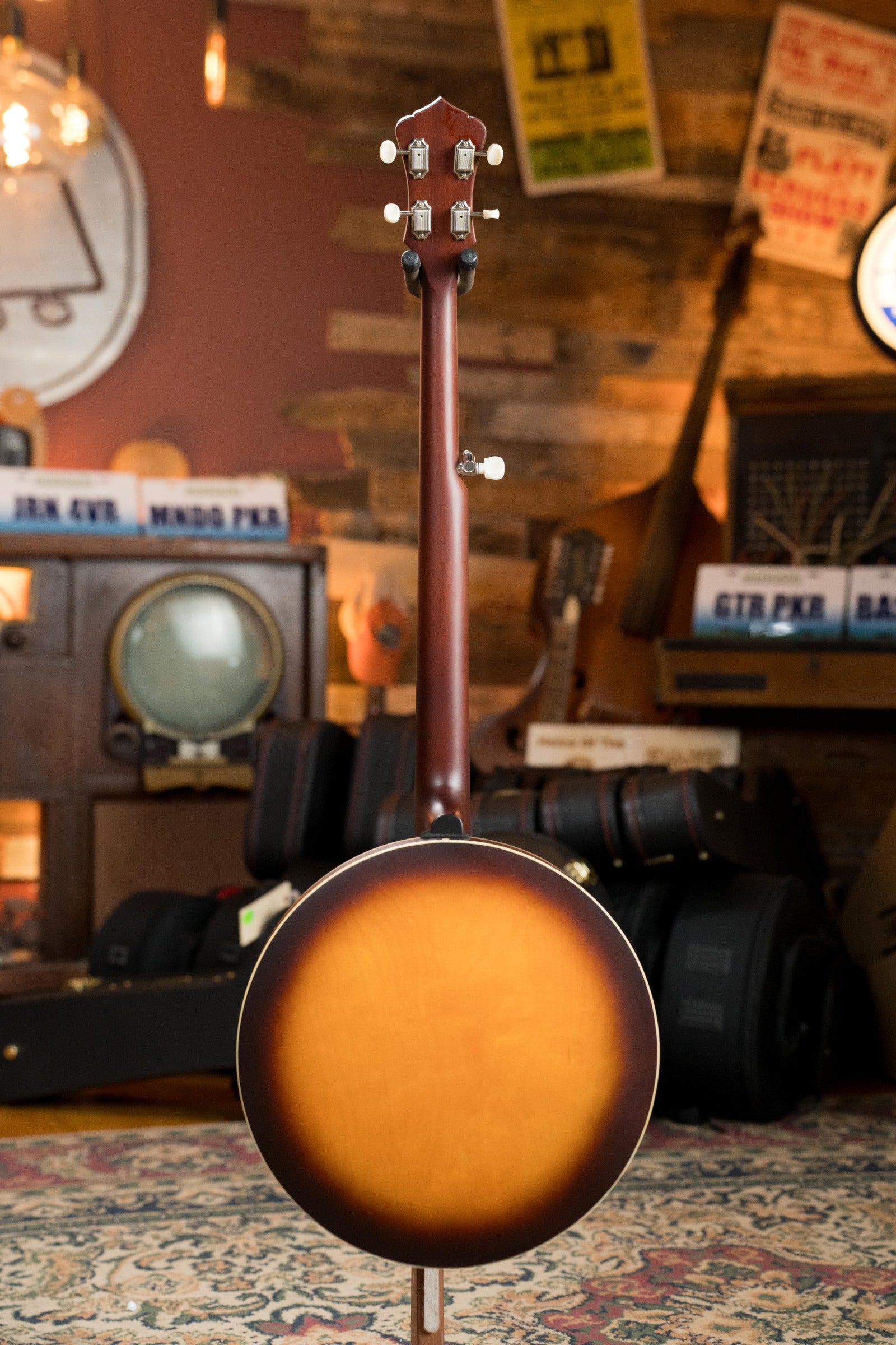 The Recording King RKH-05 Dirty 30s Resonator Banjo #2563 is displayed upright with its back facing forward in a cozy, vintage-style room filled with musical instruments, posters, and equipment cases.